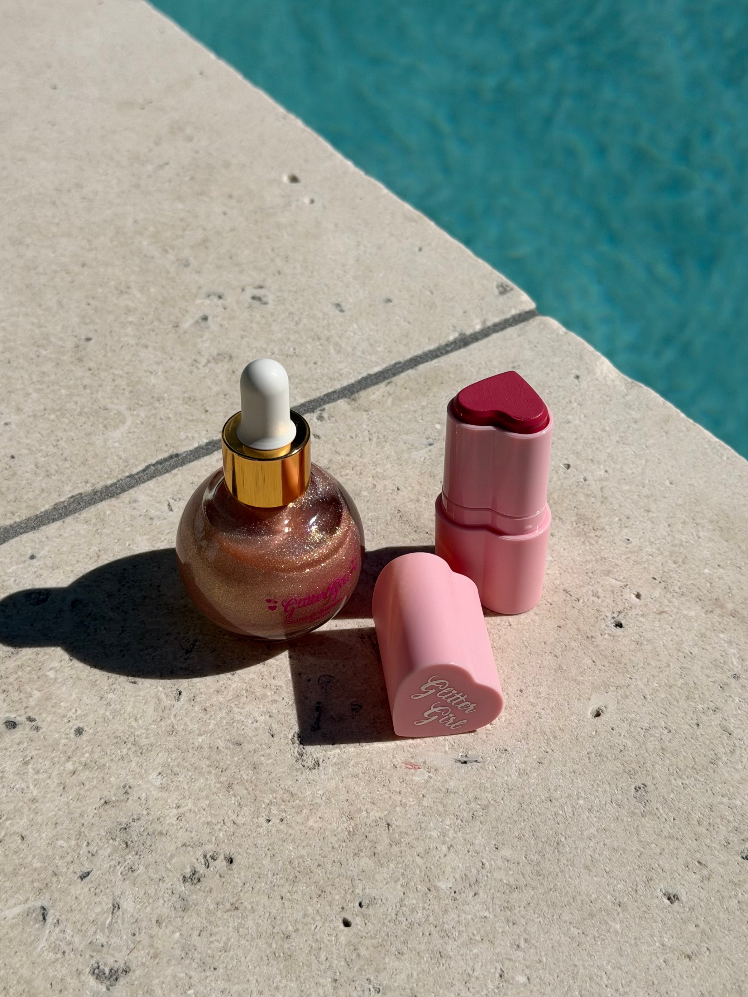A sparkling cosmetic oil and two lipsticks, one in a heart shape, placed on a light stone surface beside a pool.