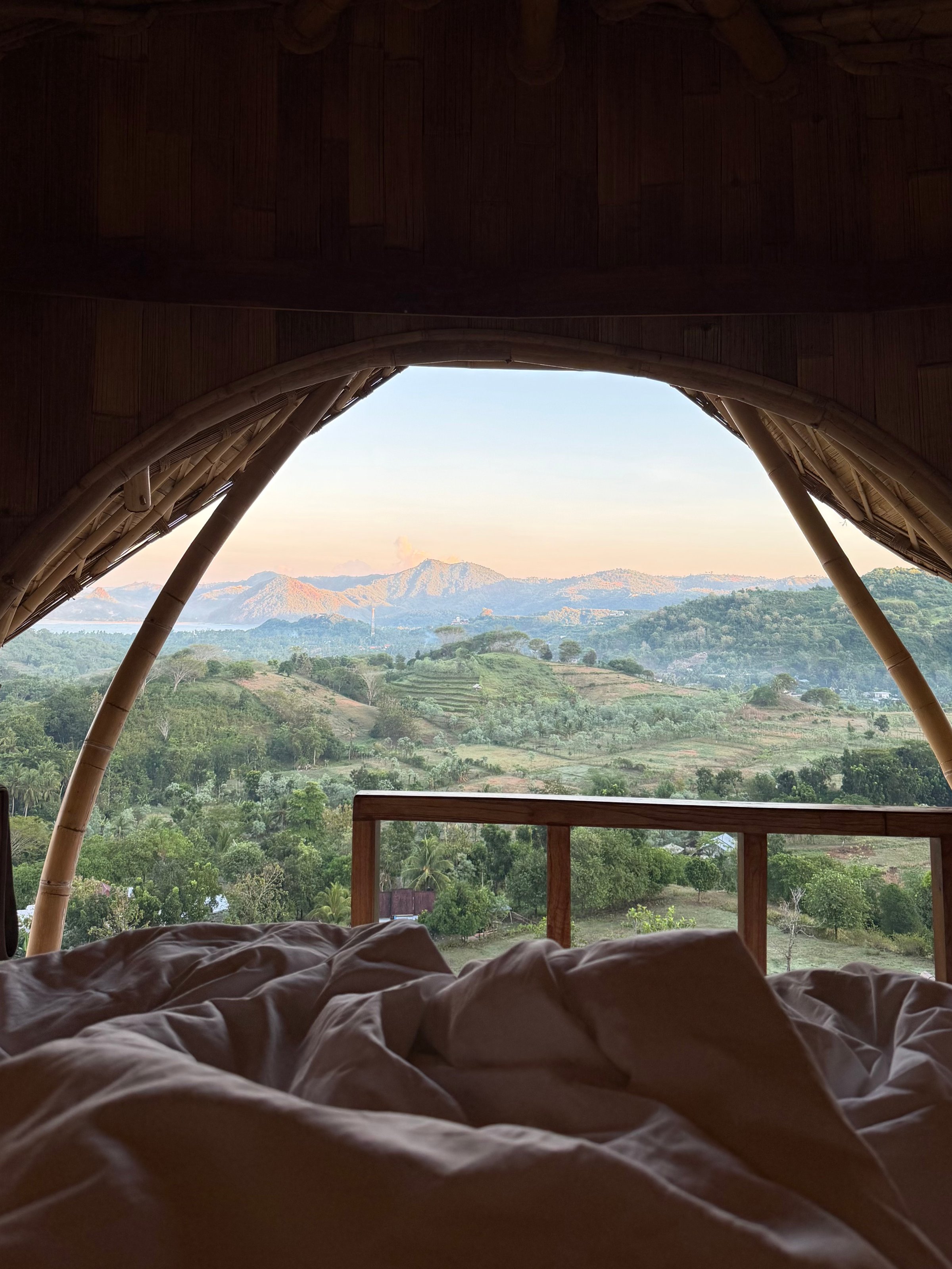 A scenic view of mountains and valleys visible through a large open window, with a cozy bed in the foreground.