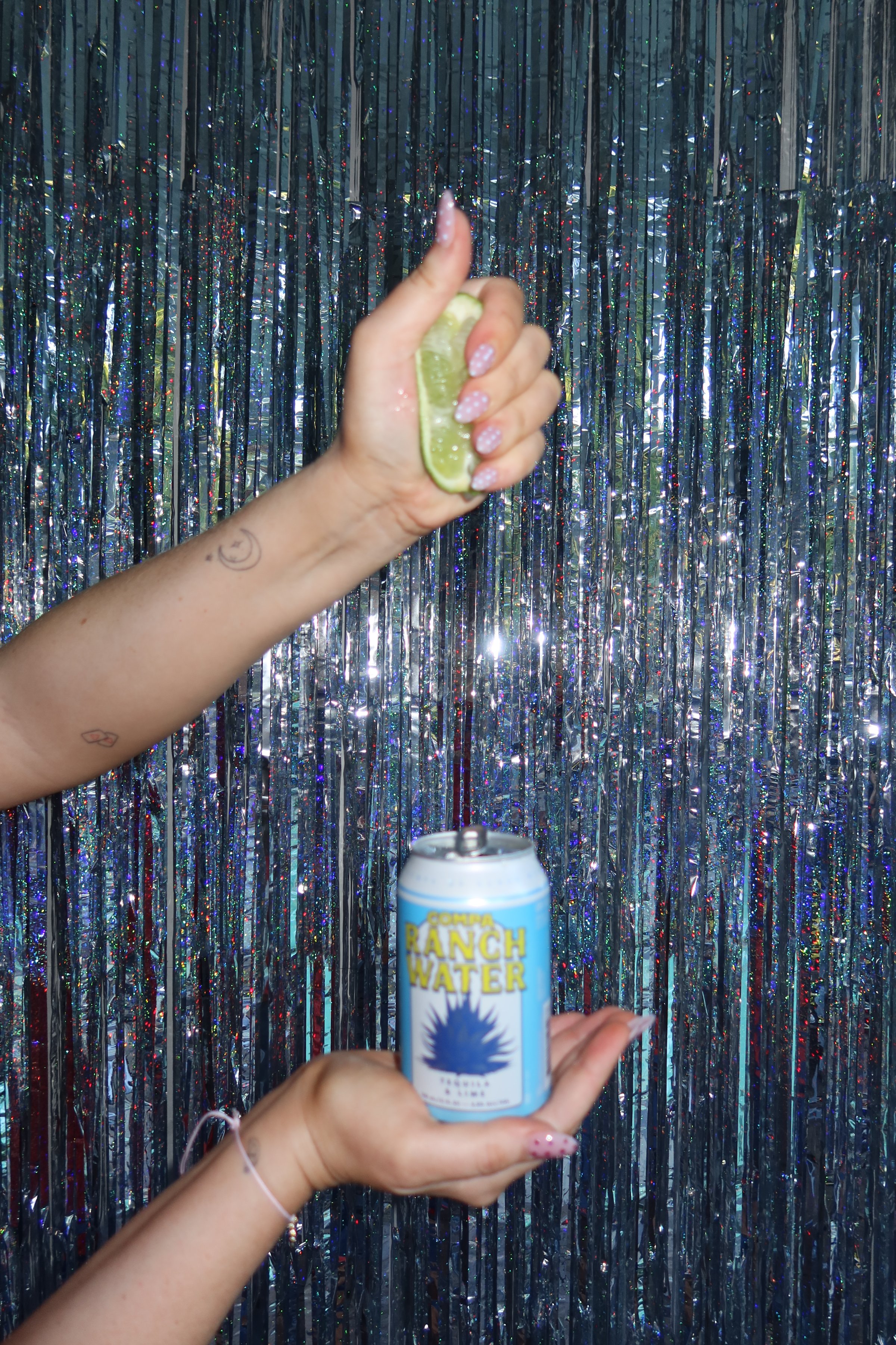 A hand squeezing a lime above a can of sparkling water, set against a shiny, metallic backdrop.