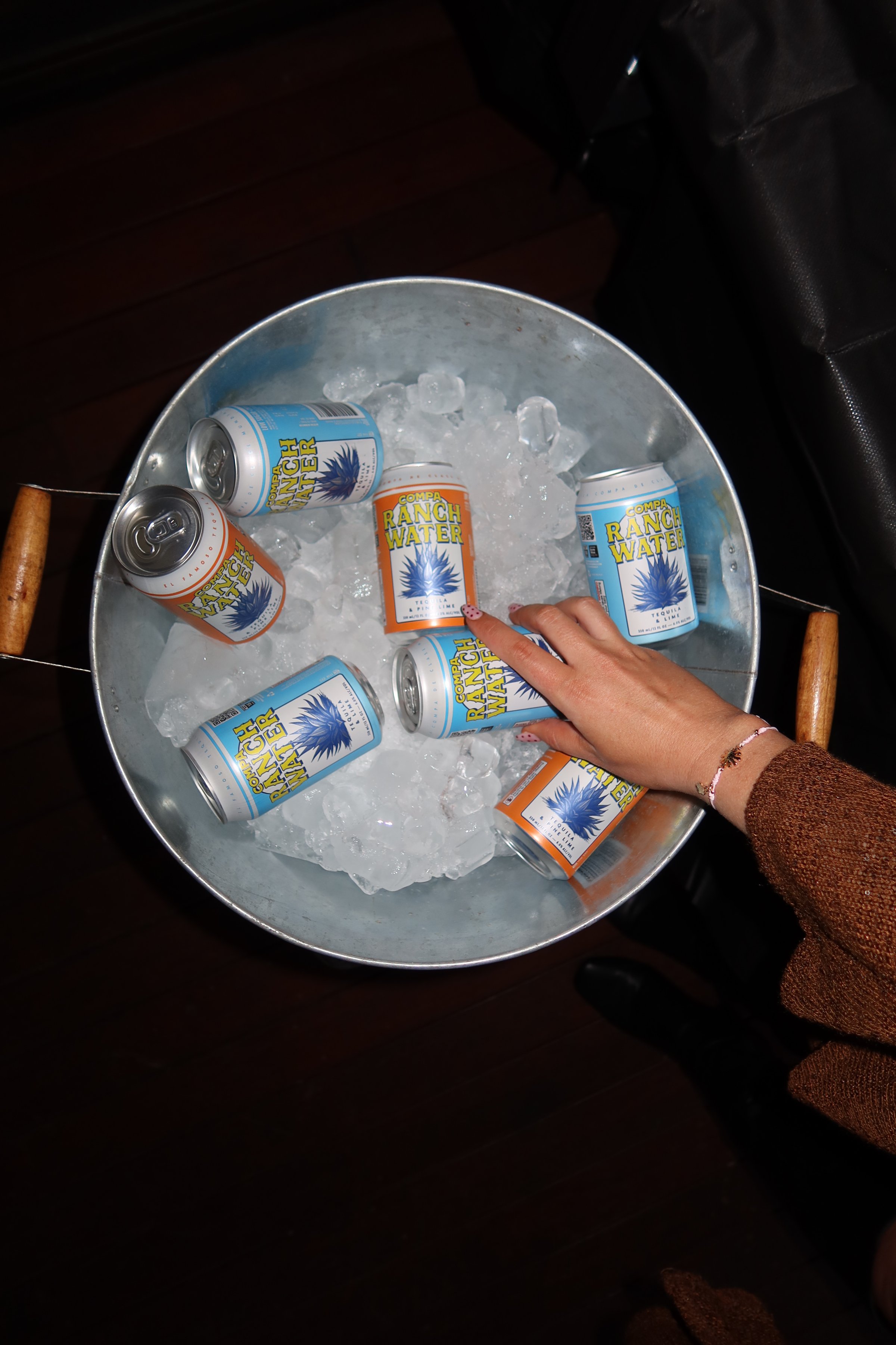 A hand reaching into a metal bucket filled with ice and colorful cans of beverage.
