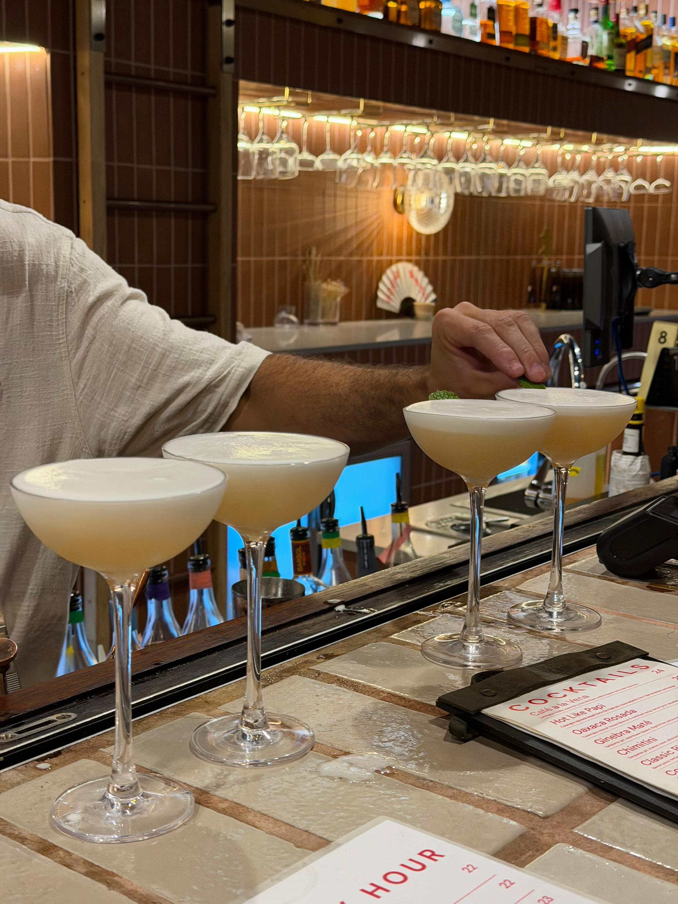 A bartender prepares cocktails at a bar, showcasing four elegant drinks in coupe glasses lined up on the counter.