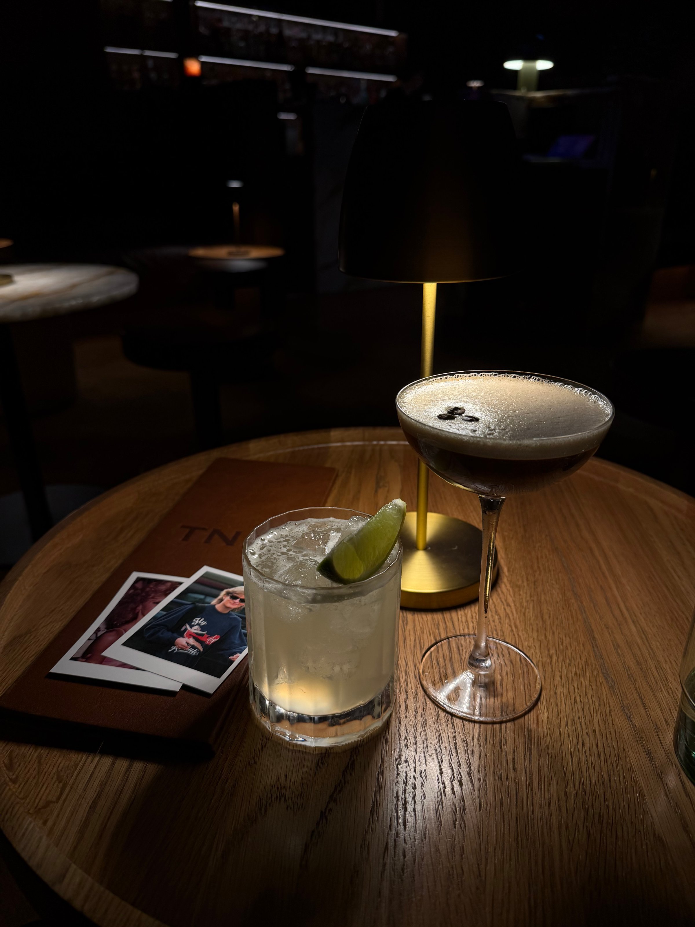 Two cocktails on a wooden table, one garnished with a lime slice and the other topped with foam, with a dimly lit background and a photo card nearby.