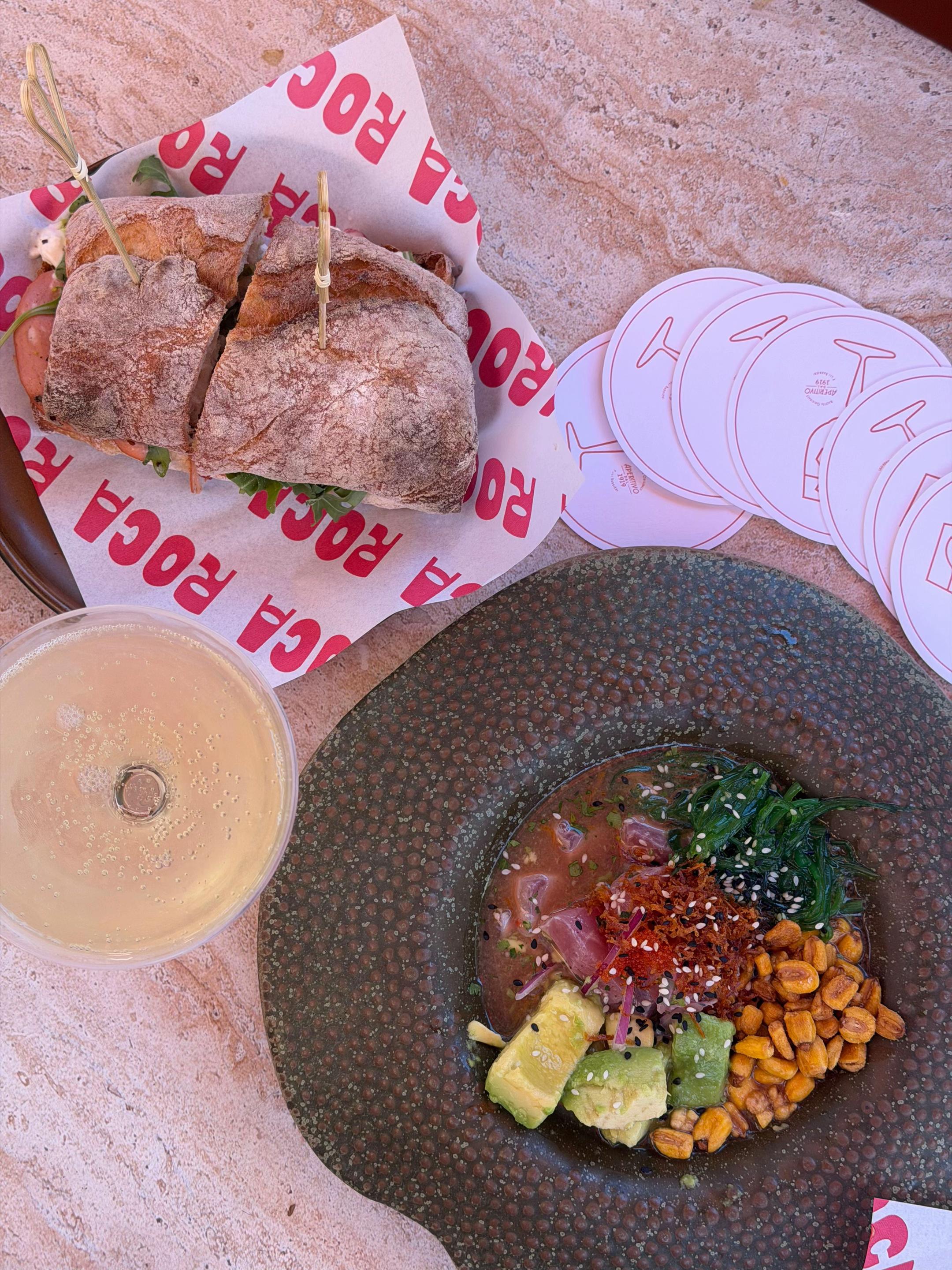 A beautifully arranged meal featuring a bowl of poke with fresh ingredients and a sandwich on a branded paper. A drink is also visible in the foreground.