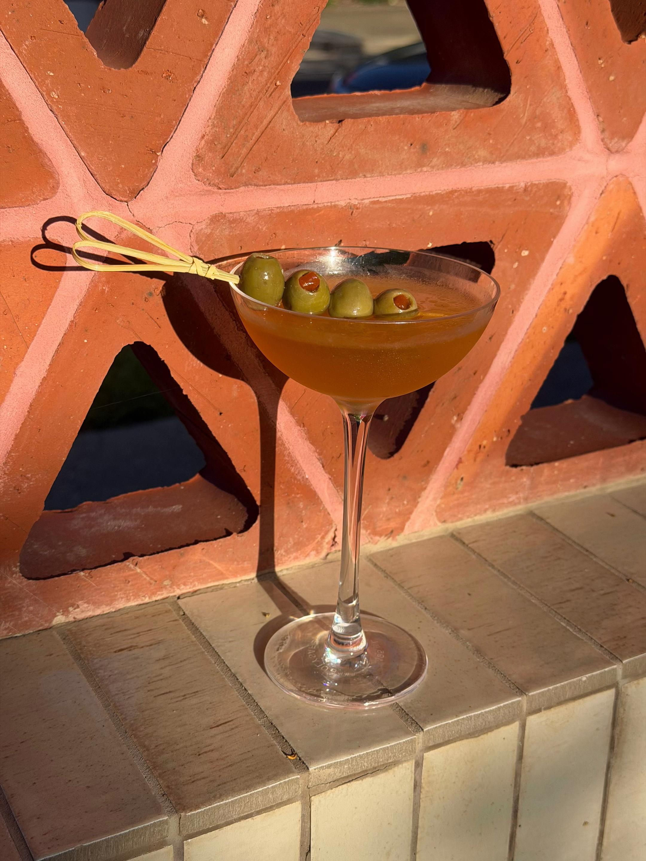 A cocktail served in a elegant glass, garnished with olives, placed on a tiled surface against a textured wall.
