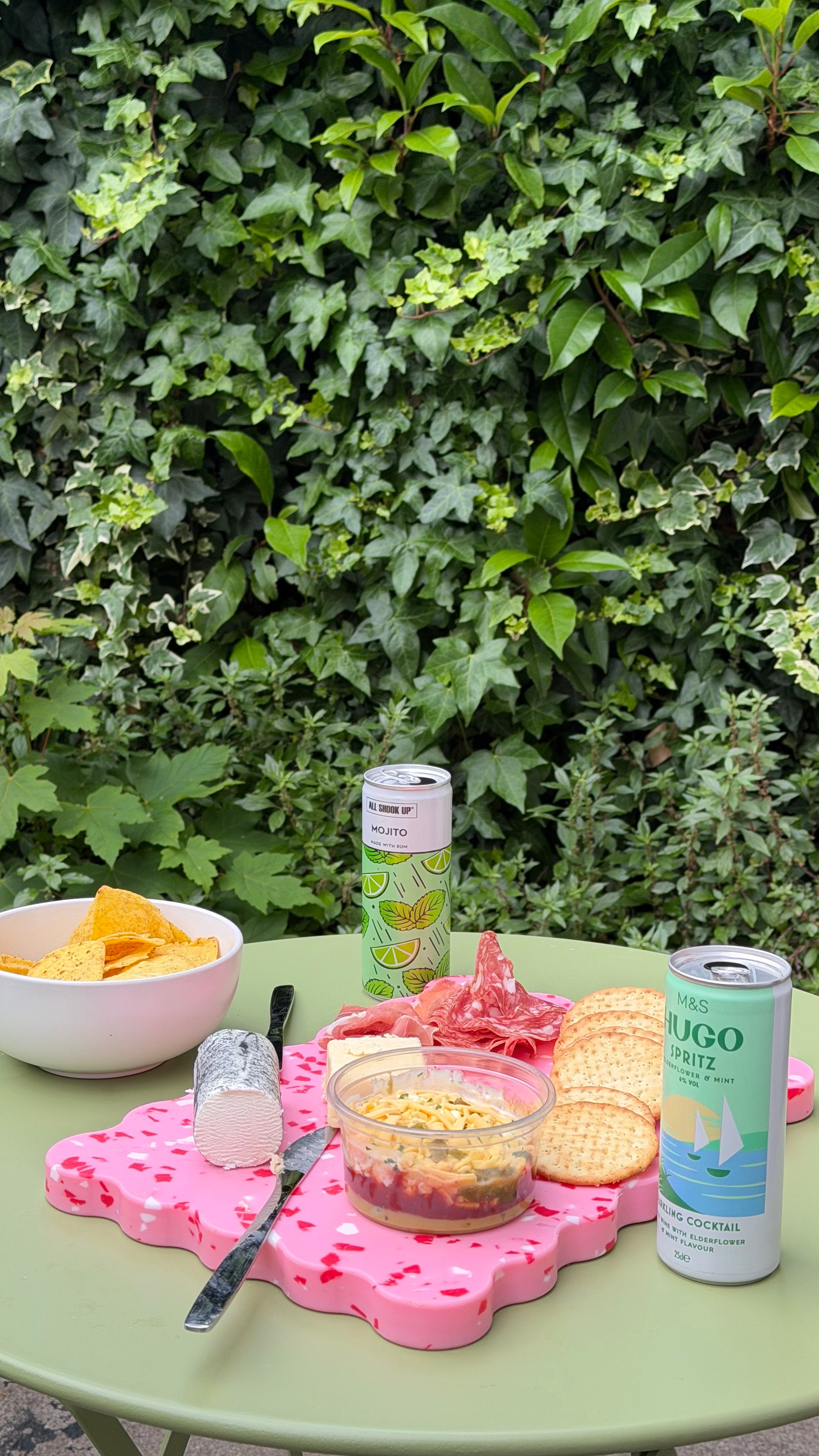 A vibrant outdoor table setting featuring a bowl of chips, a platter with cheese, crackers, and cured meats, alongside two cans of sparkling beverages, all set against a lush green backdrop.