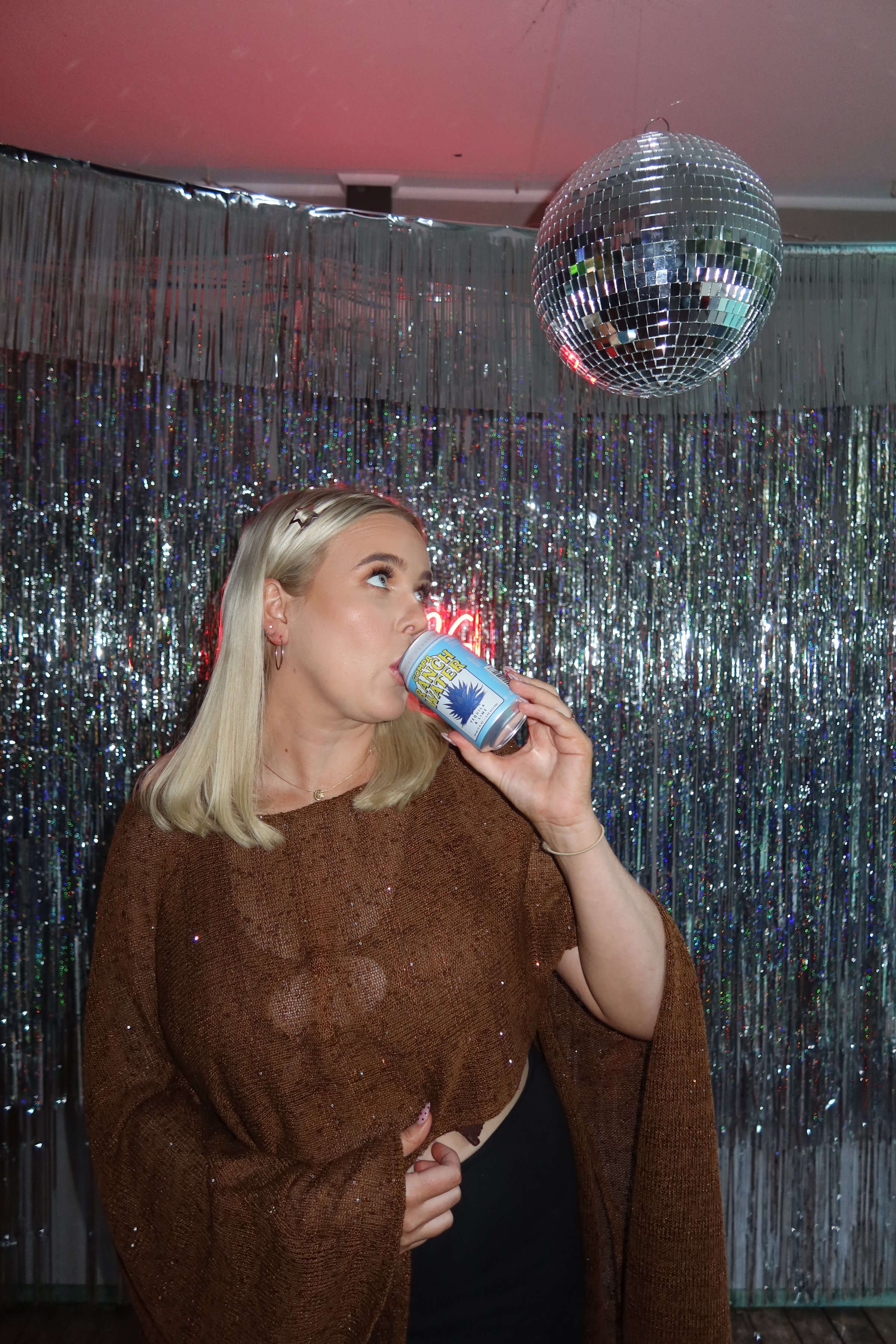A woman in a sparkly brown top drinks from a can while looking up at a disco ball, surrounded by a shimmering backdrop.
