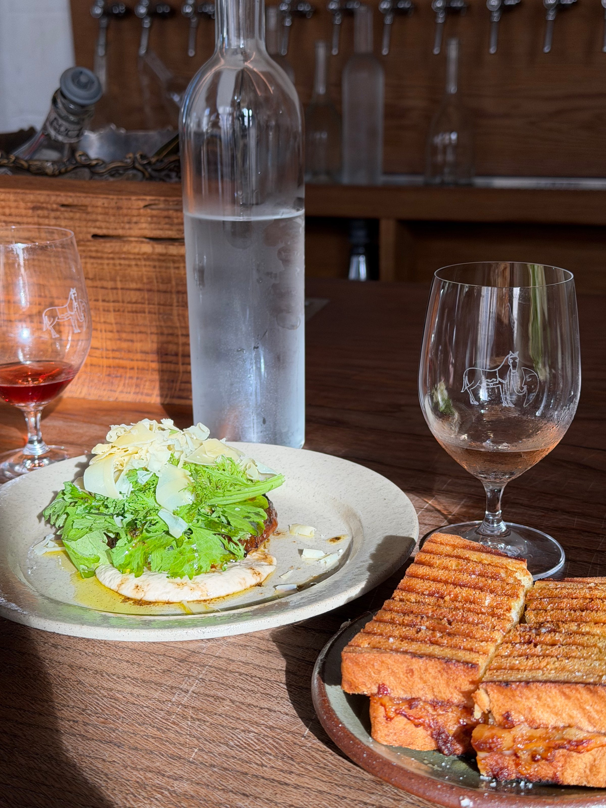 A beautifully arranged plate featuring a sandwich with greens and a side of crispy toast, accompanied by glasses of water and wine on a wooden table.