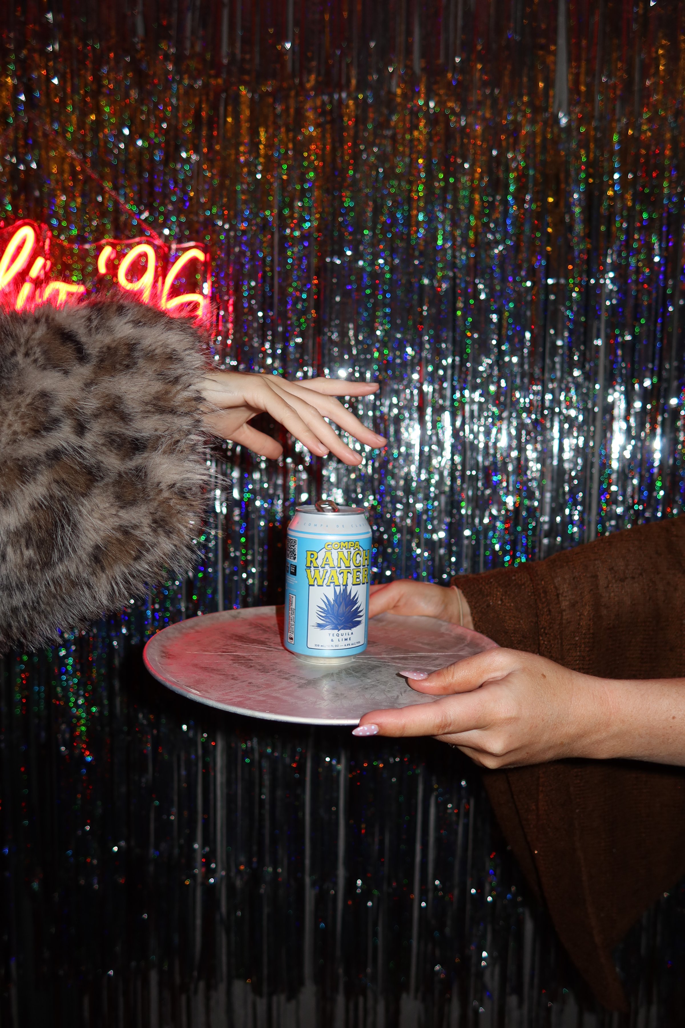 A hand in a furry coat reaches for a can of drink being offered on a plate by another hand, set against a sparkling backdrop.