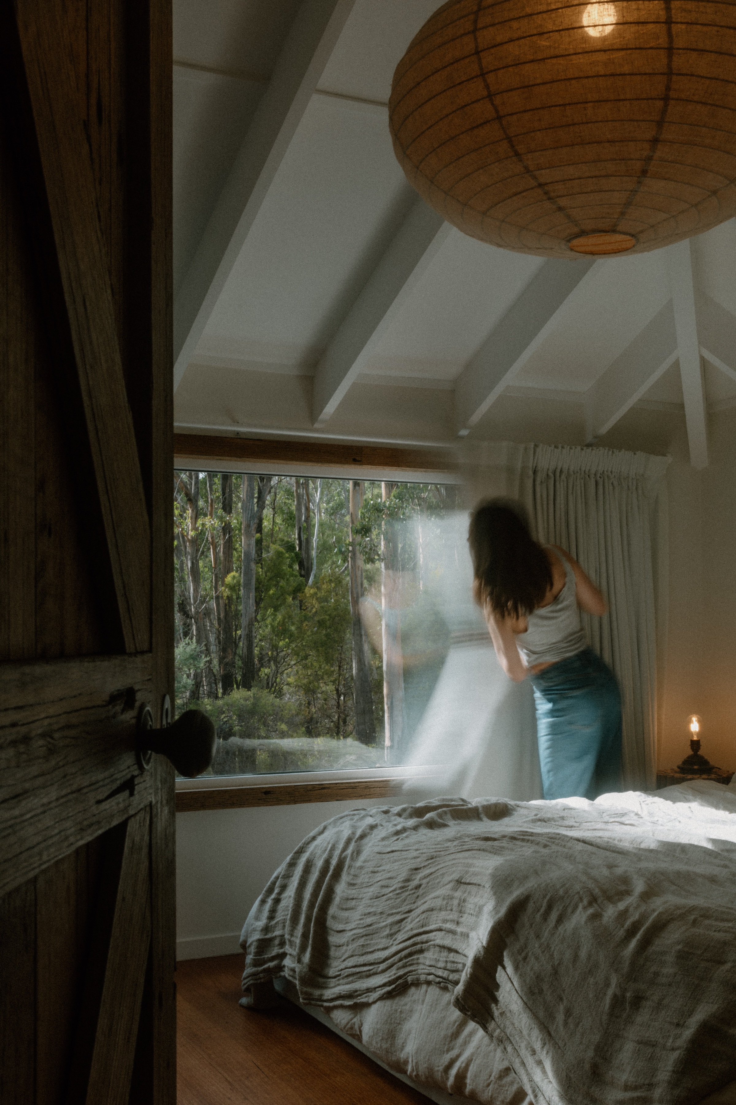 A person stands by a window in a cozy bedroom, pulling back sheer curtains to reveal a view of trees outside. The room features a bed with a textured blanket and a warm ambiance from a nearby candle.