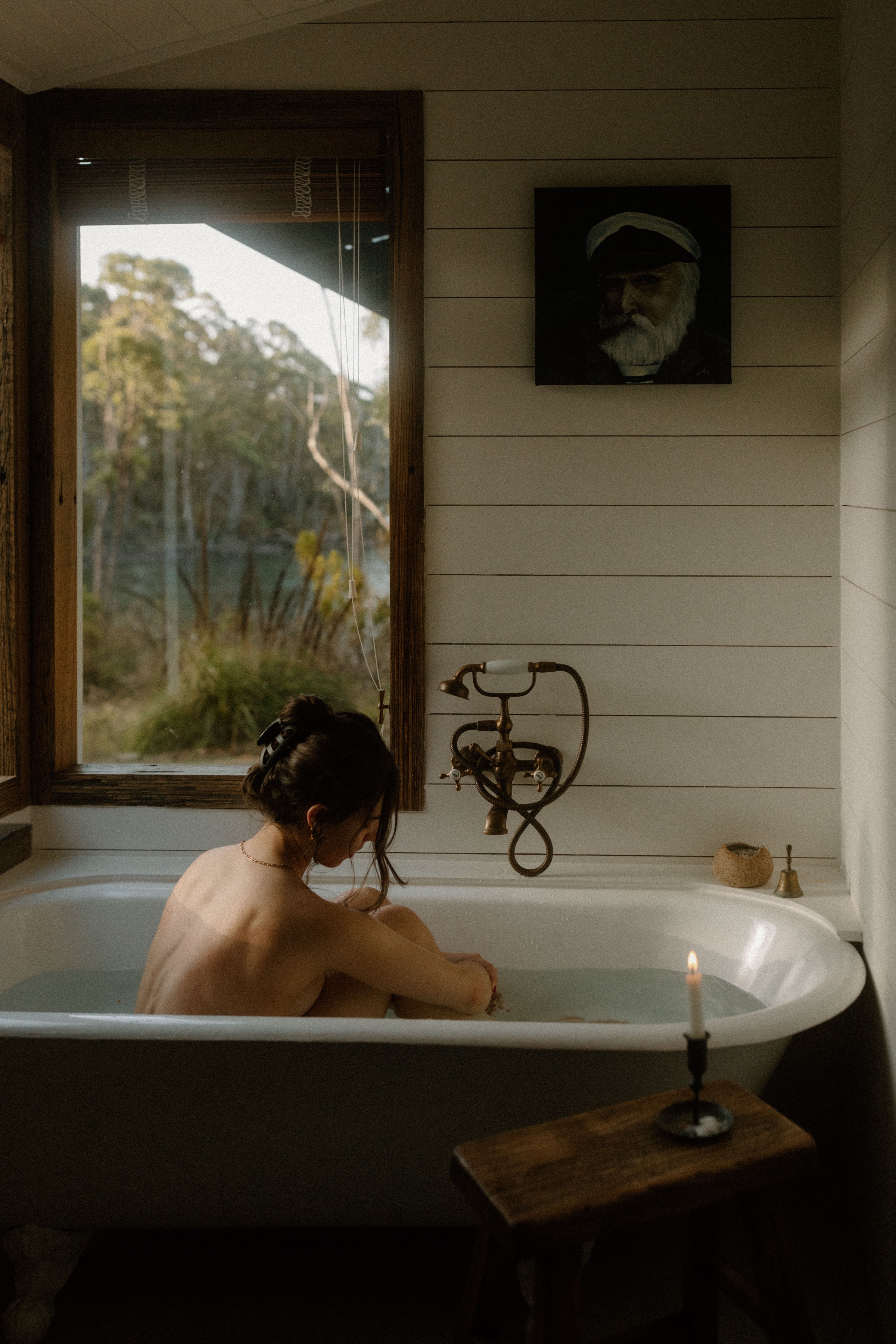 A person sitting in a vintage bathtub, gazing out a window with natural light illuminating the scene. The setting has a rustic feel with wooden accents and a candle on a nearby table.