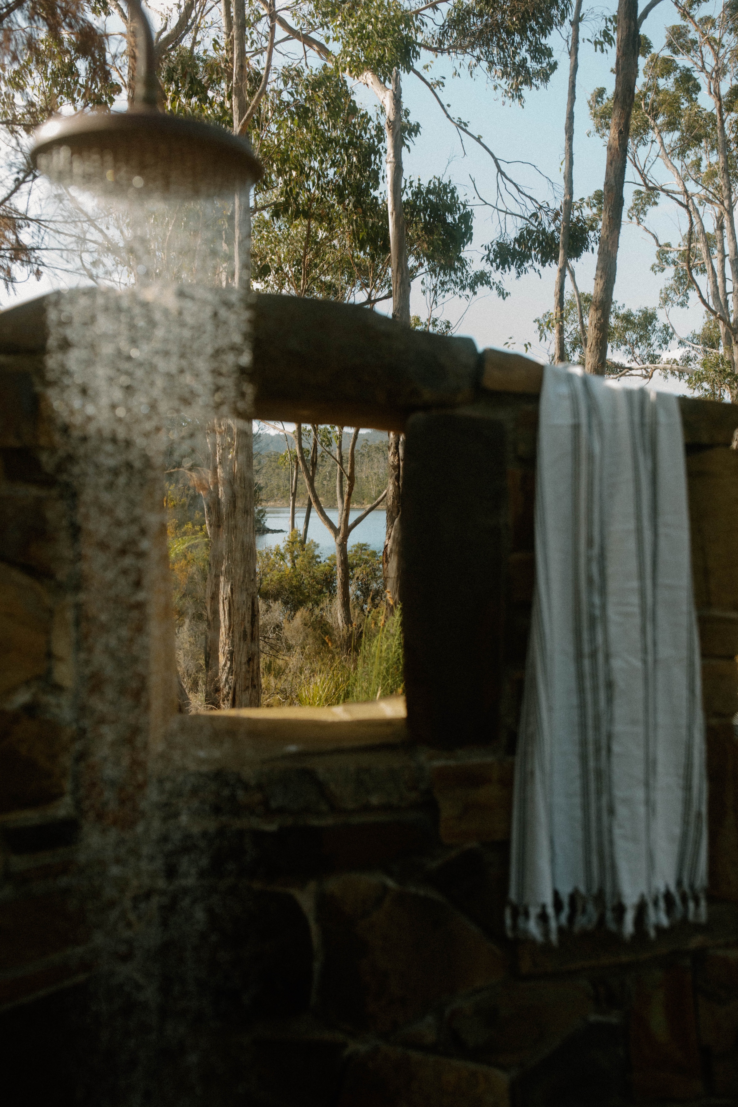A serene outdoor shower surrounded by lush greenery, with a view of a lake in the background. A white towel hangs nearby, adding a touch of comfort to the natural setting.