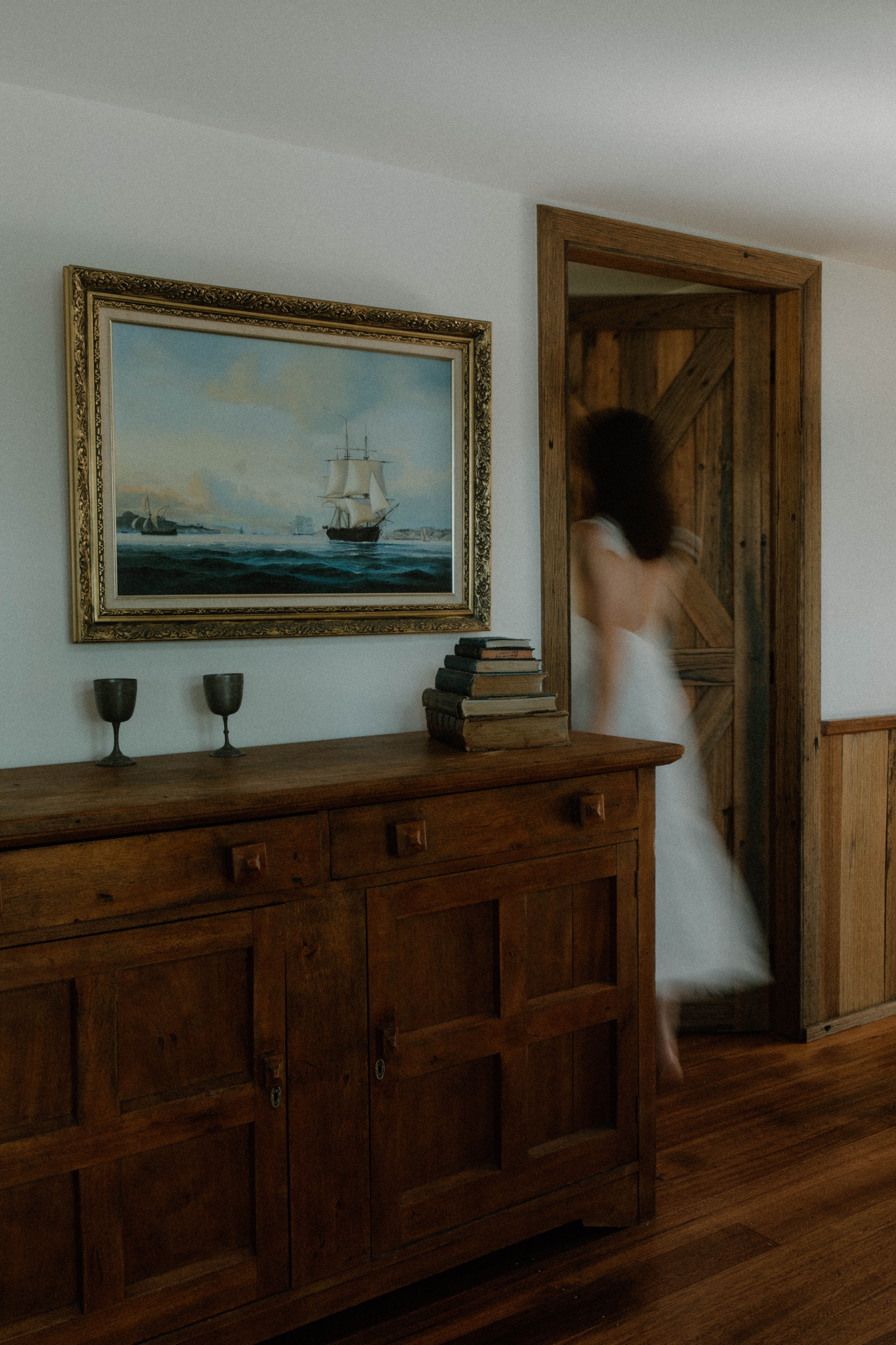 A blurred figure in a white dress walks past a wooden door in a room featuring a vintage ship painting and a wooden sideboard with stacked books and two candlesticks.