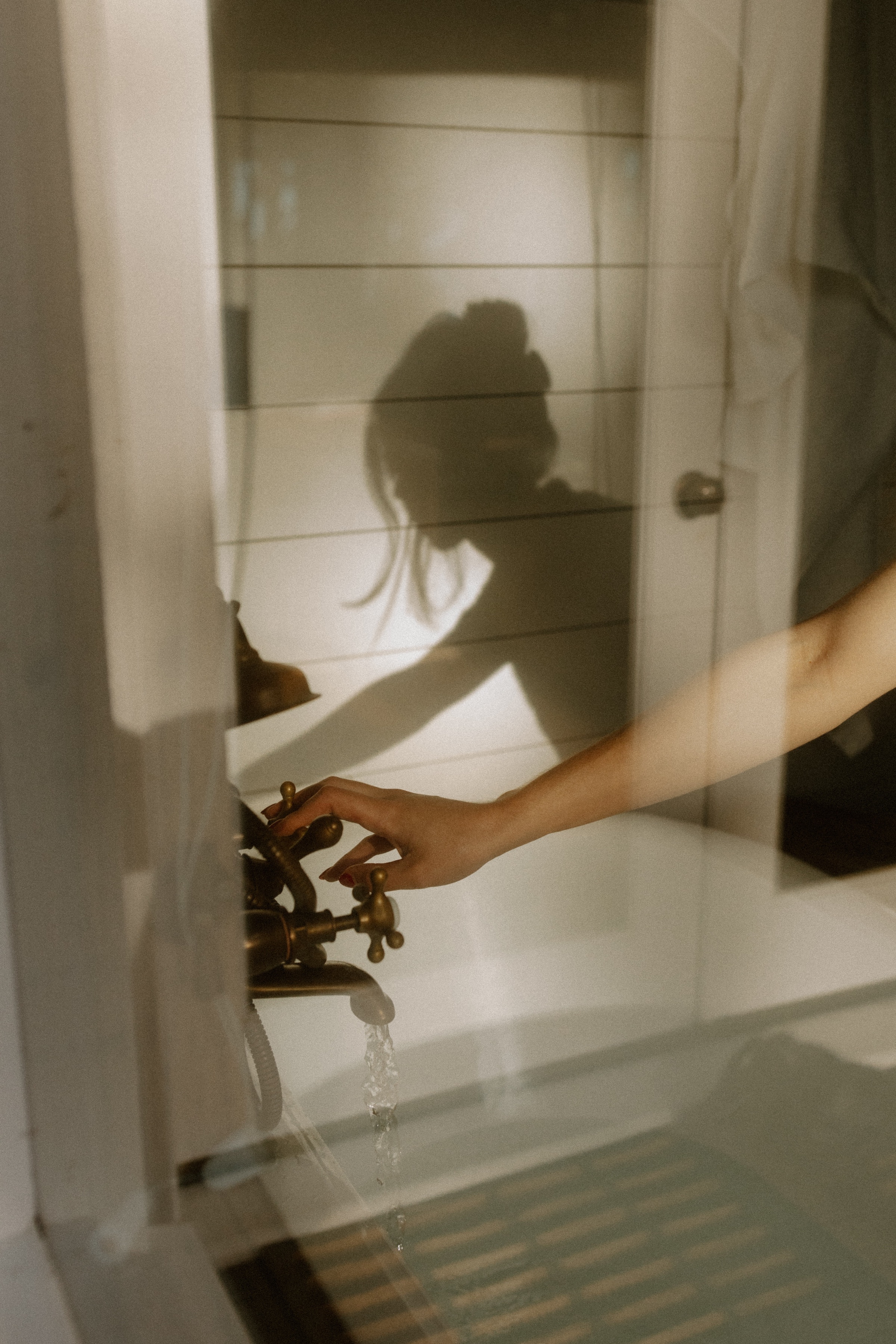 A hand reaching for a vintage faucet, with a soft shadow cast on the wall behind. The scene is illuminated by natural light, creating a serene atmosphere.
