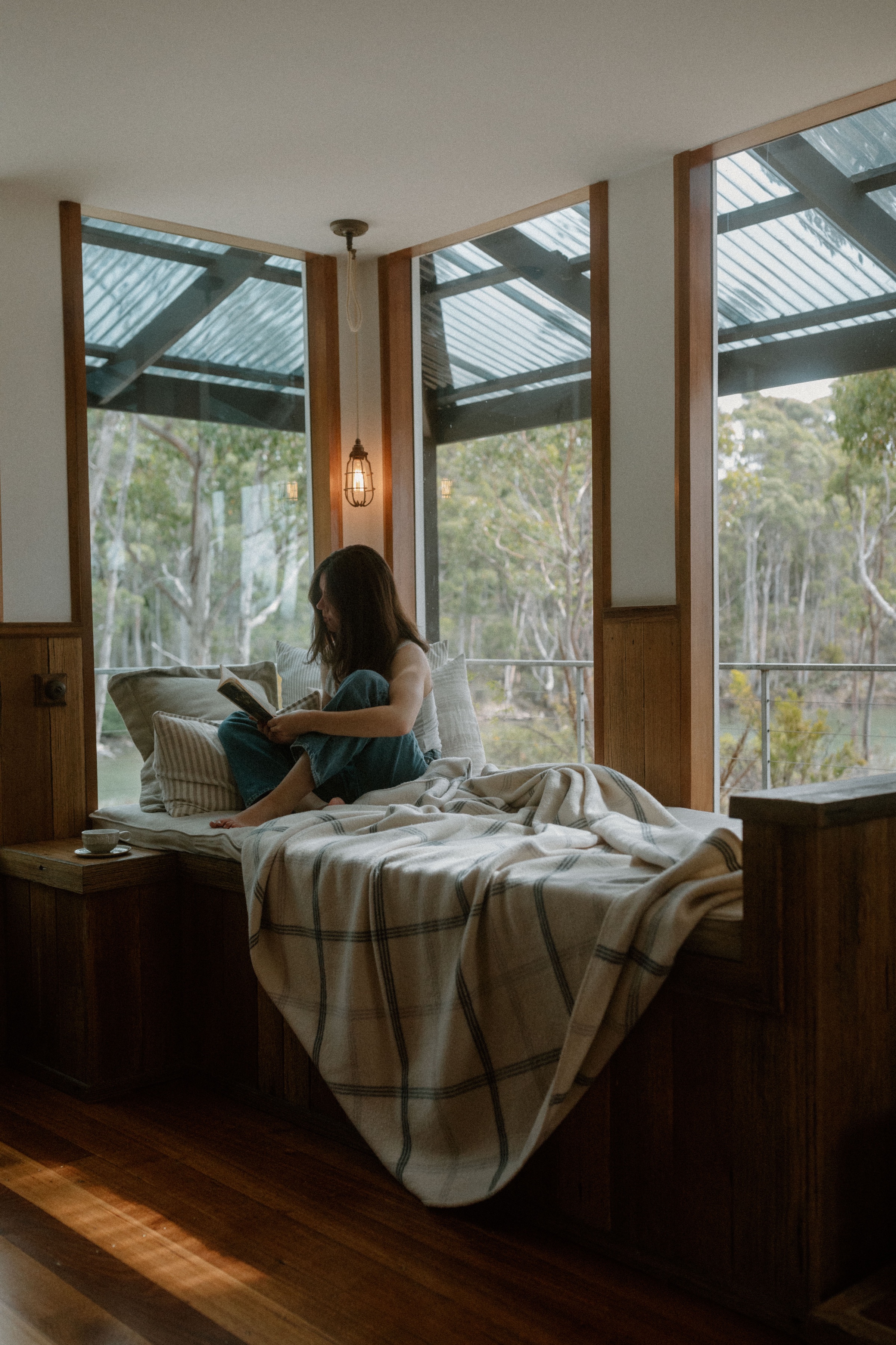 A person sitting on a bed in a cozy room, reading a book with large windows showcasing a natural landscape.