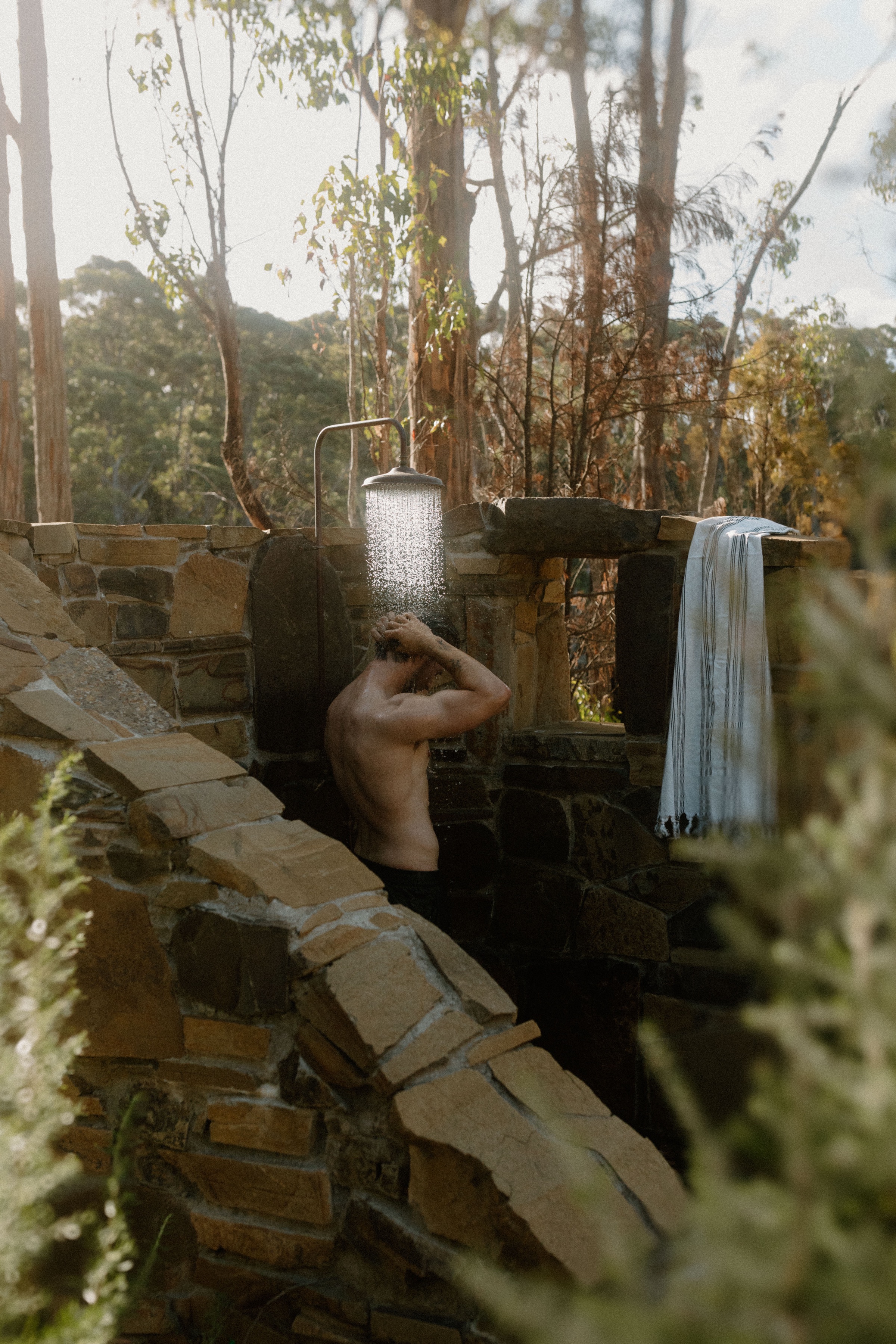 A person showering outdoors in a rustic stone shower surrounded by greenery and trees.
