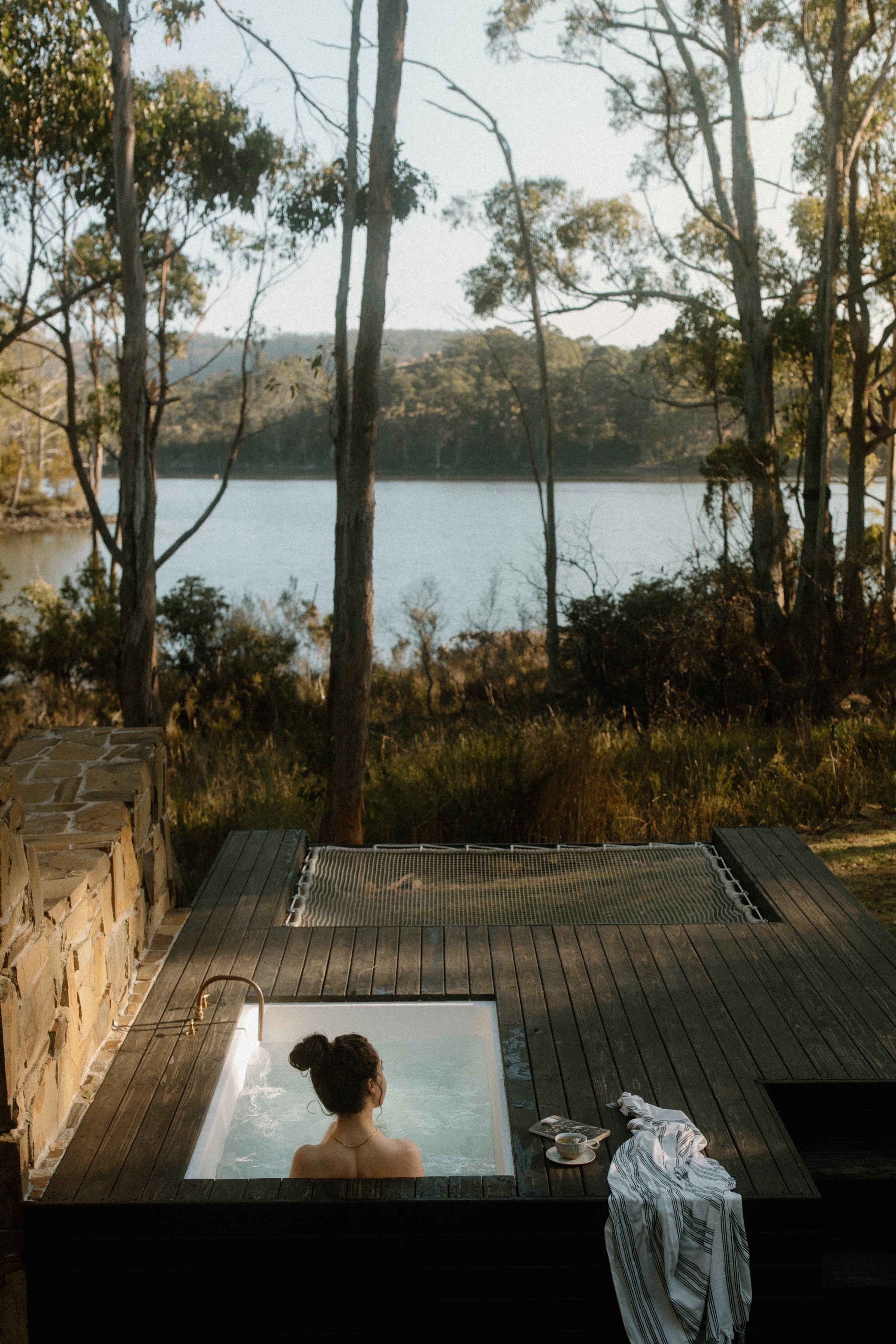 A person relaxing in a bathtub on a wooden deck overlooking a serene lake surrounded by trees.