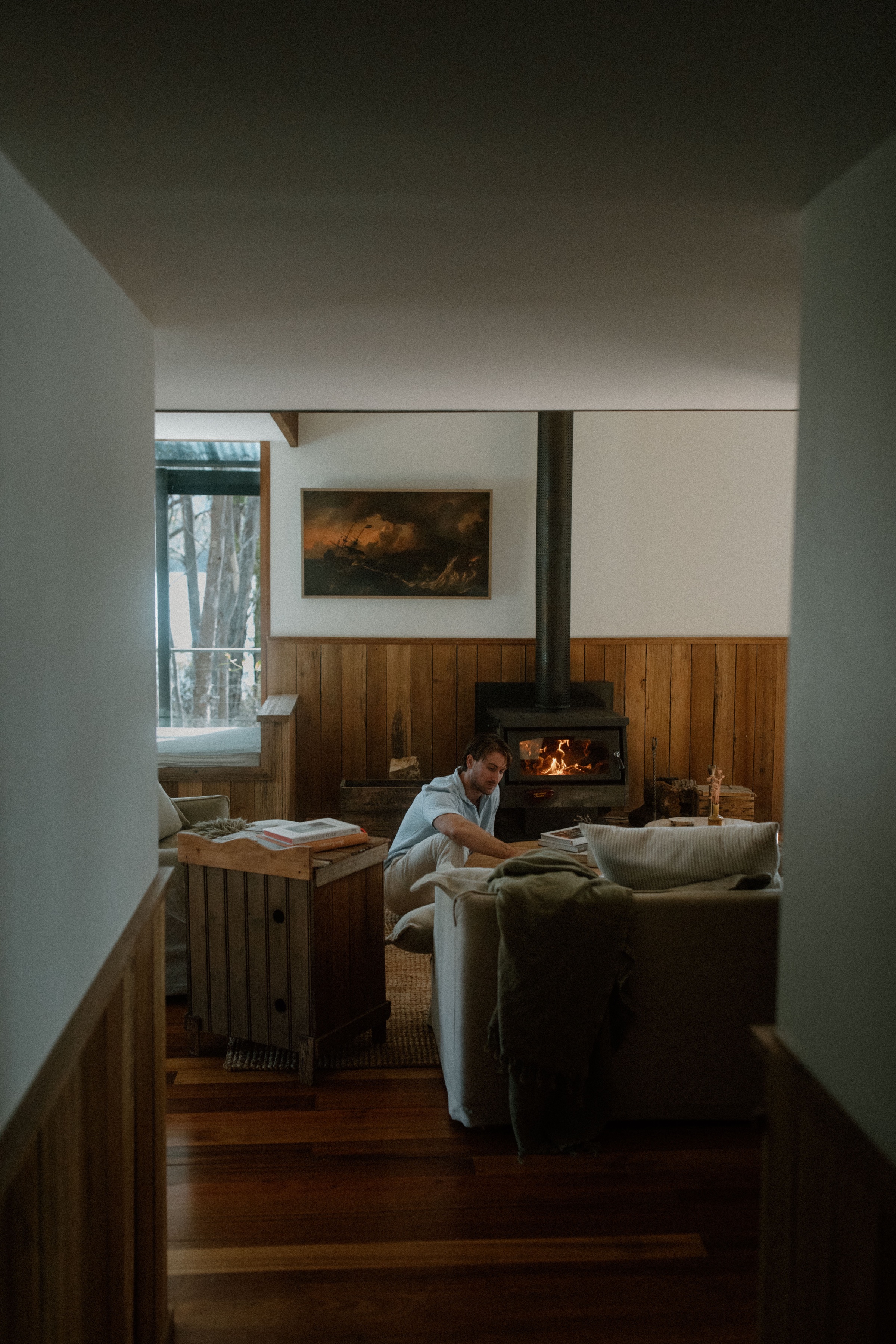 A cozy living room scene with a person sitting comfortably by a fireplace, surrounded by wooden decor and soft furnishings.