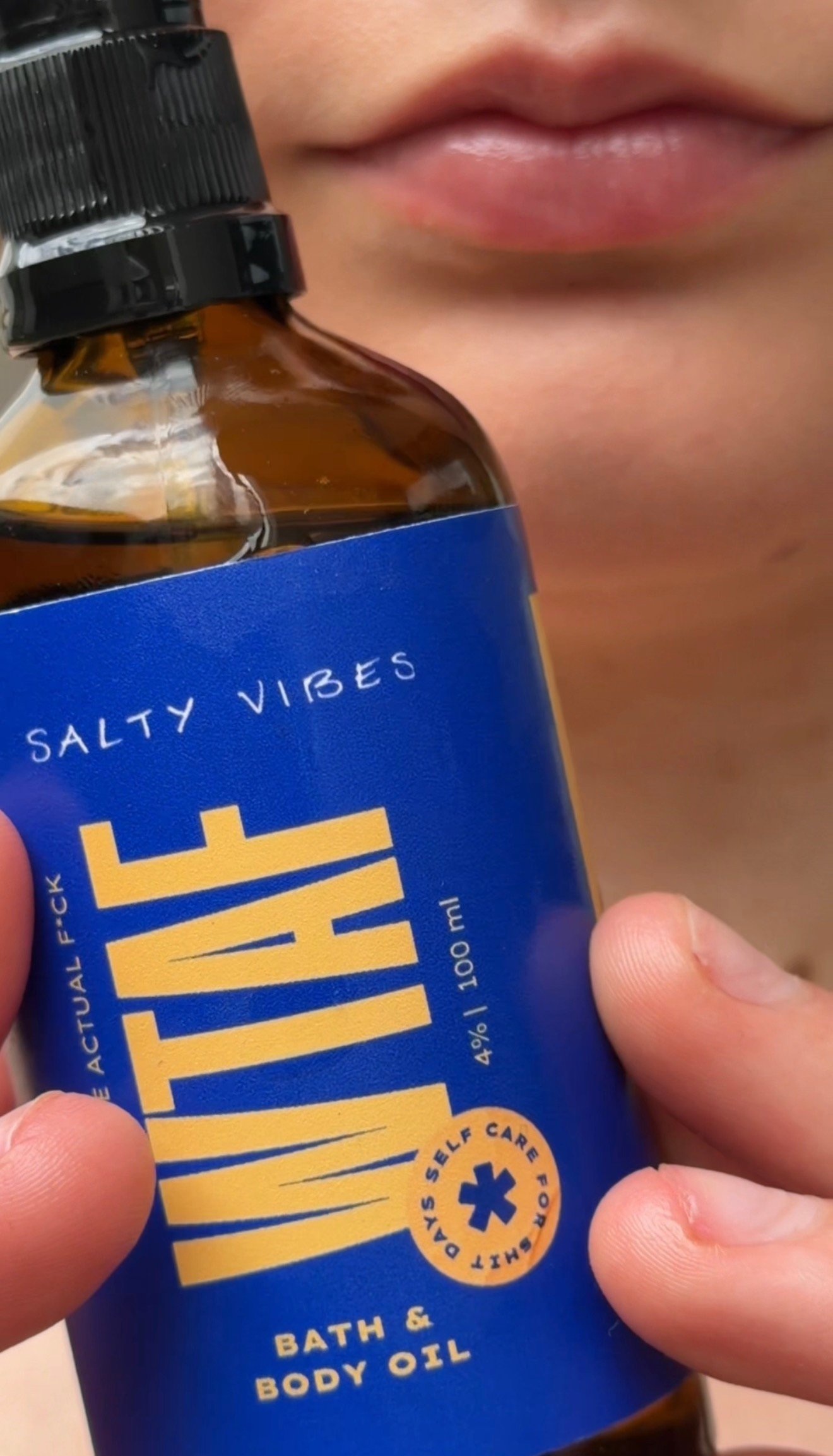 A close-up of a person holding a bottle of bath and body oil labeled 'WTAF Salty Vibes'. The bottle is dark brown with a blue label featuring bold text.