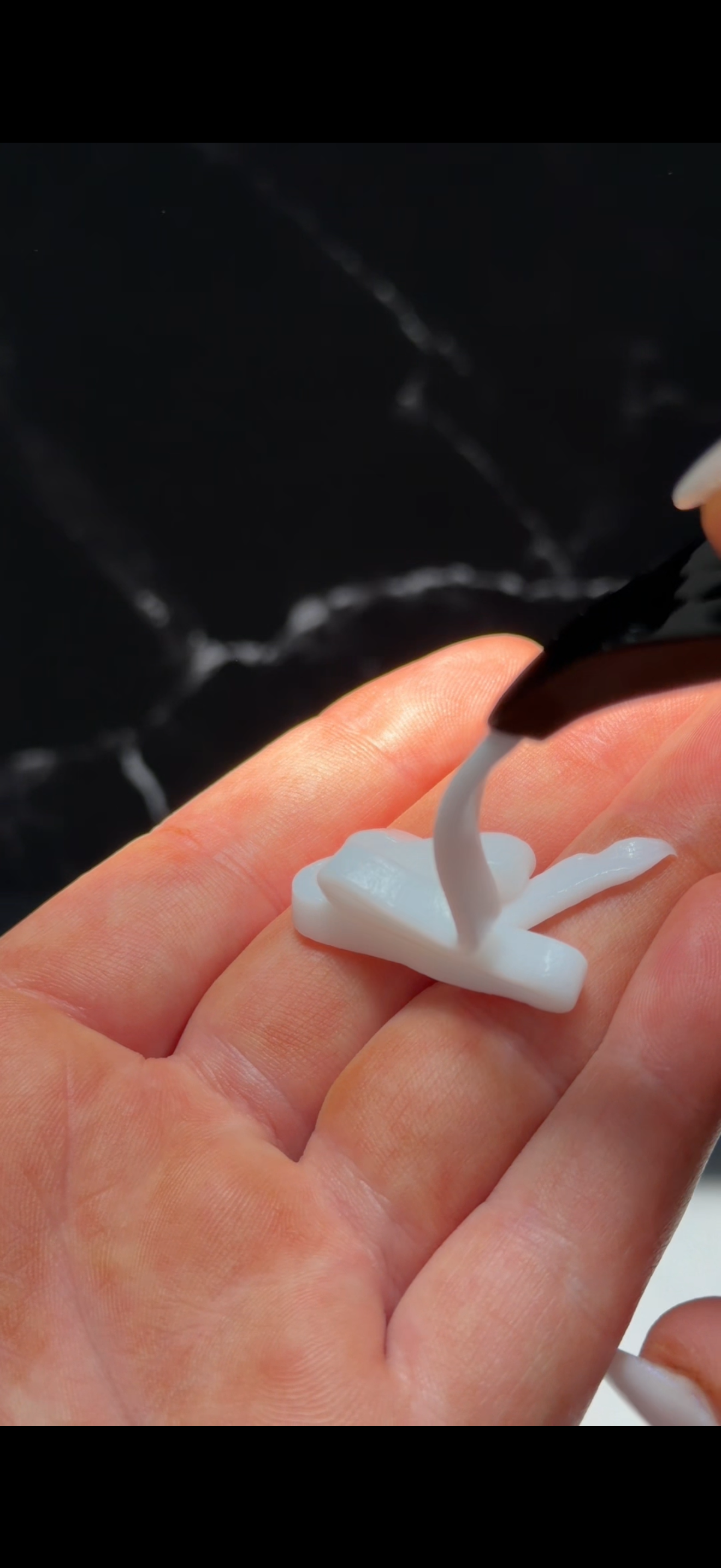 A close-up of a hand holding a small white object, with a black tool applying a substance onto it, set against a dark marble background.