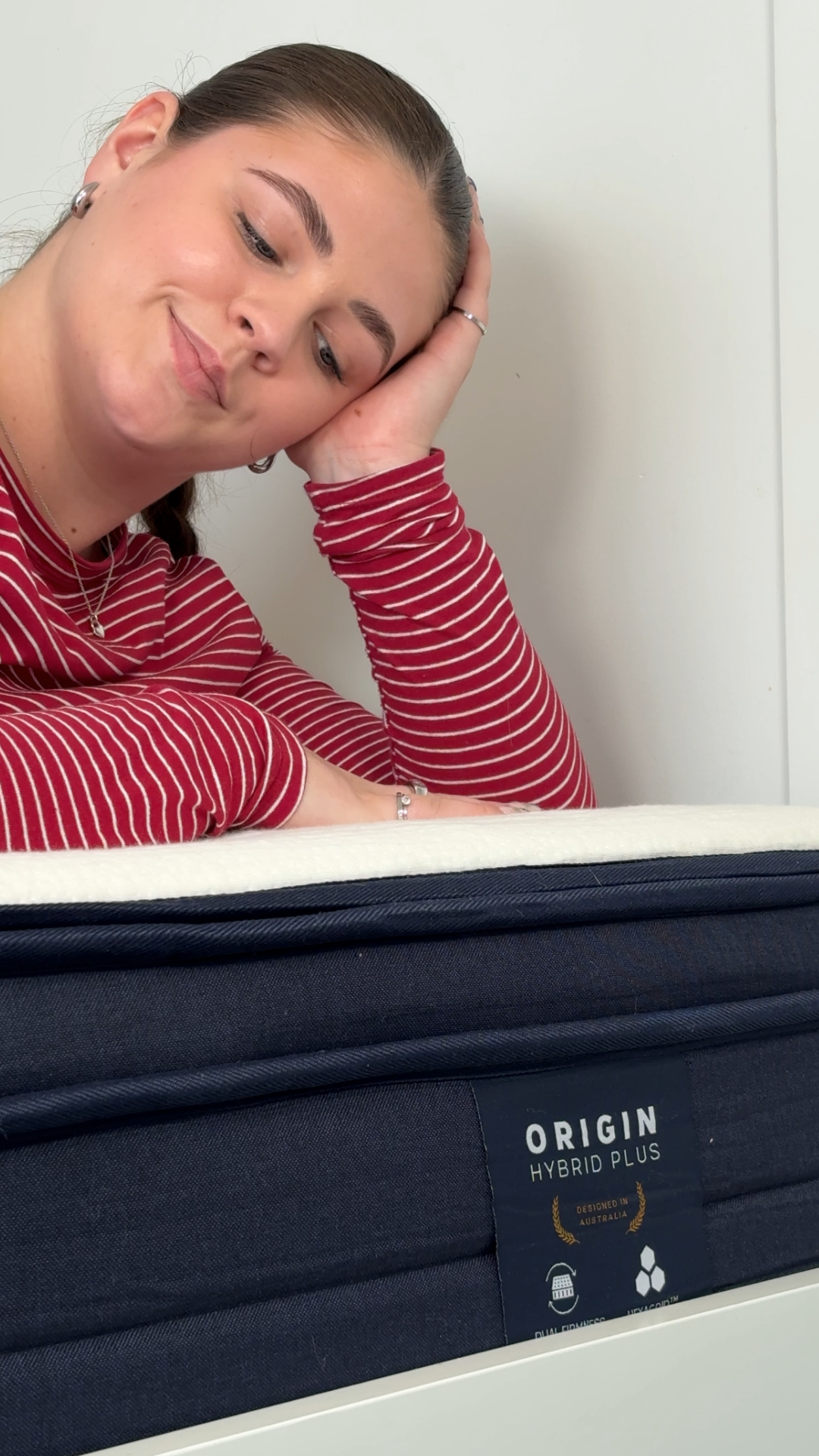A woman in a red striped shirt rests her head on her hand while looking at a mattress labeled 'Origin Hybrid Plus'.
