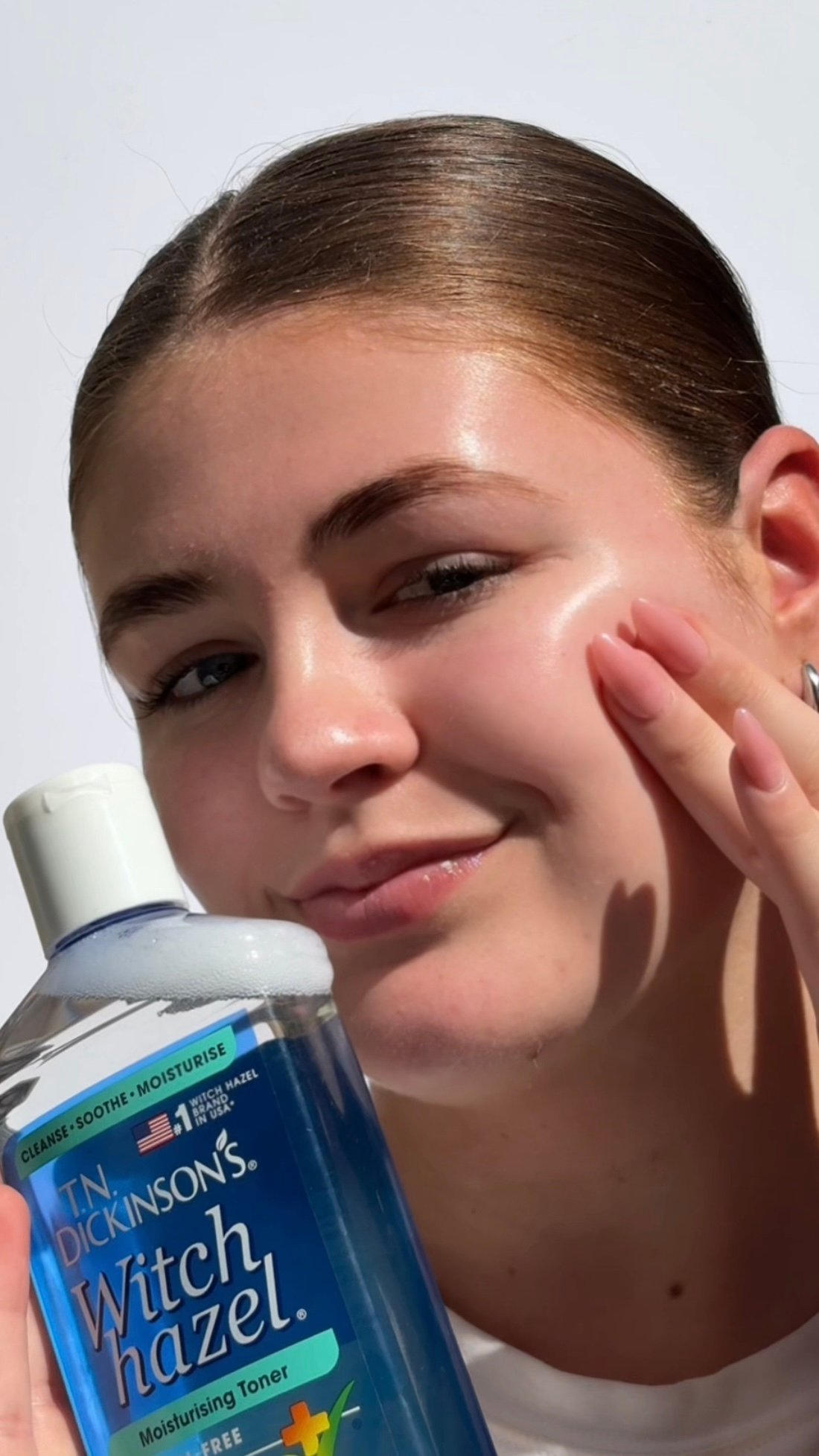 A young woman applies toner to her face, showcasing a clear and glowing complexion. She holds a bottle of T.N. Dickinson's Witch Hazel in her hand.