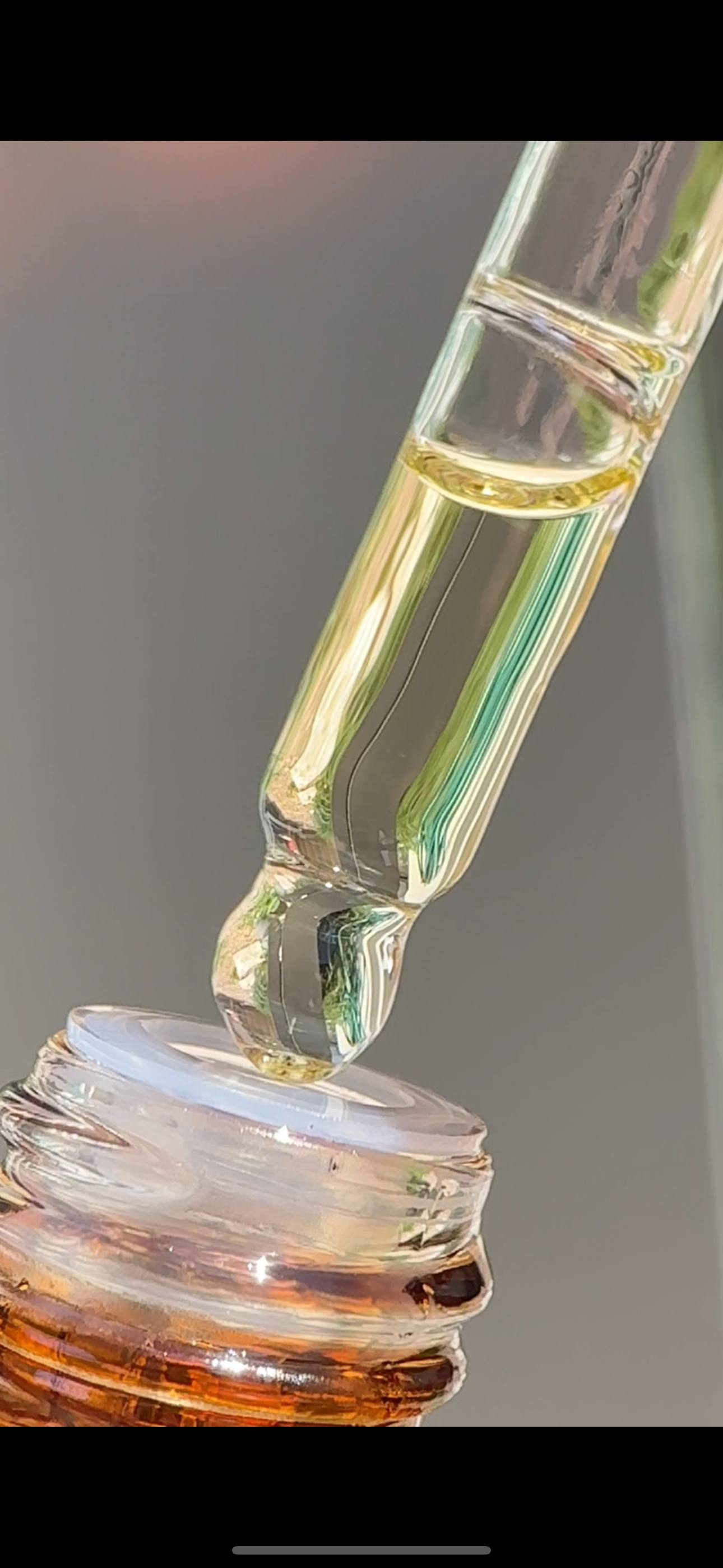 A close-up of a dropper releasing a clear liquid onto the opening of a small amber glass bottle.