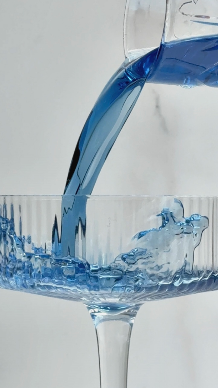 A close-up shot of blue liquid being poured into a clear glass, creating ripples in the glass.