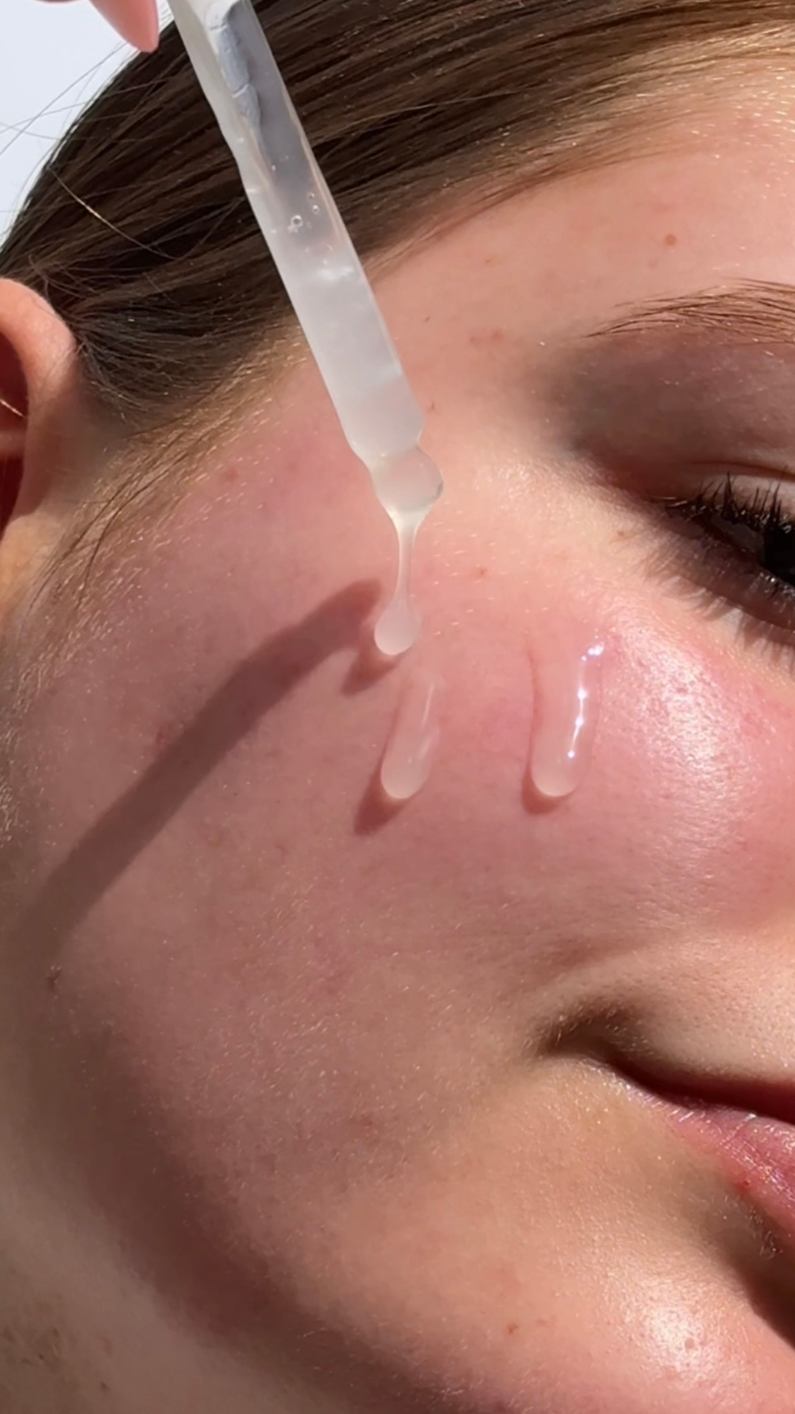 A close-up of a person's face with a dropper applying a clear serum to their cheek, highlighting skin texture and glow.