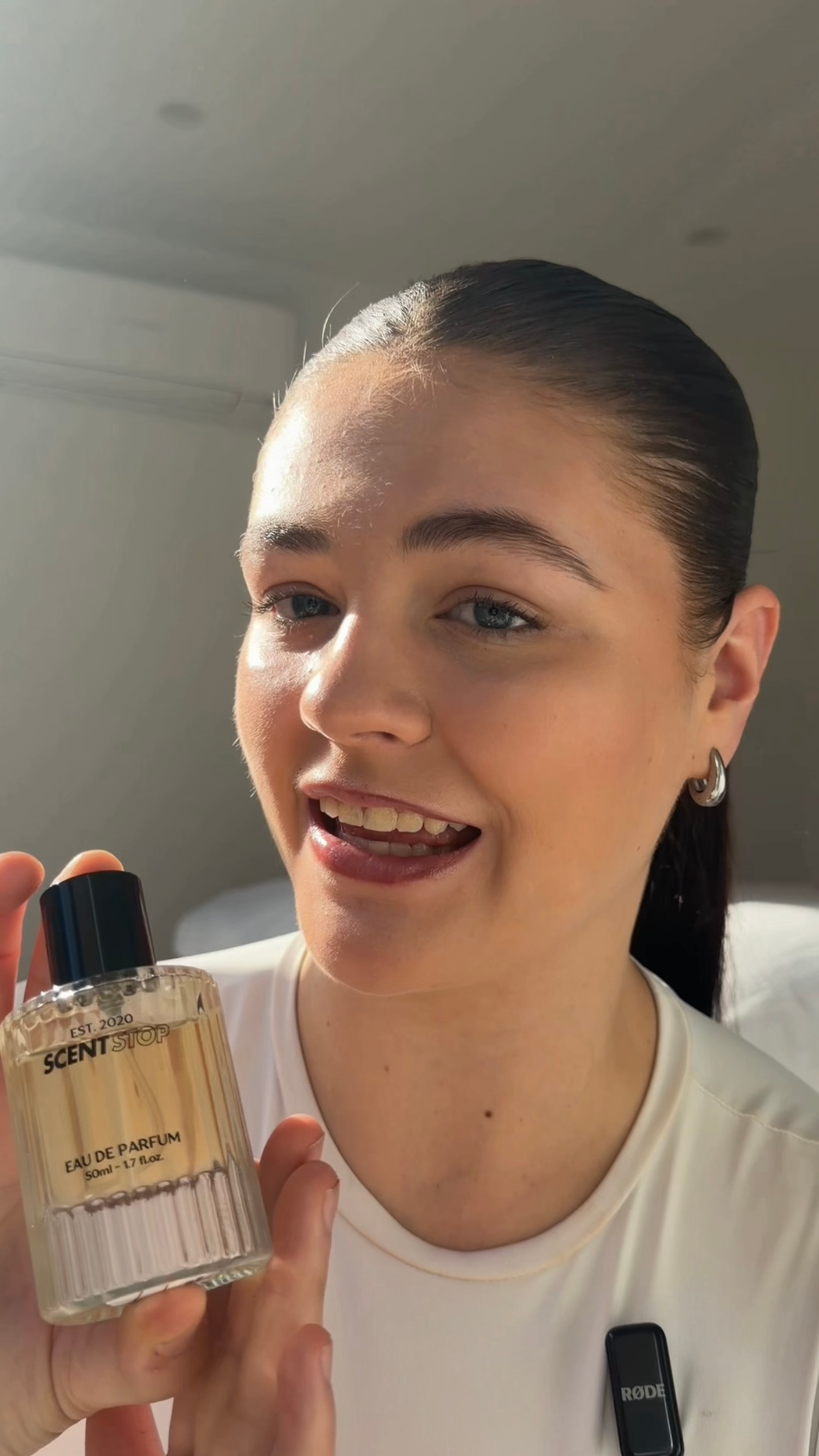 A young woman holds a bottle of perfume, smiling and showcasing the product in a well-lit indoor setting.