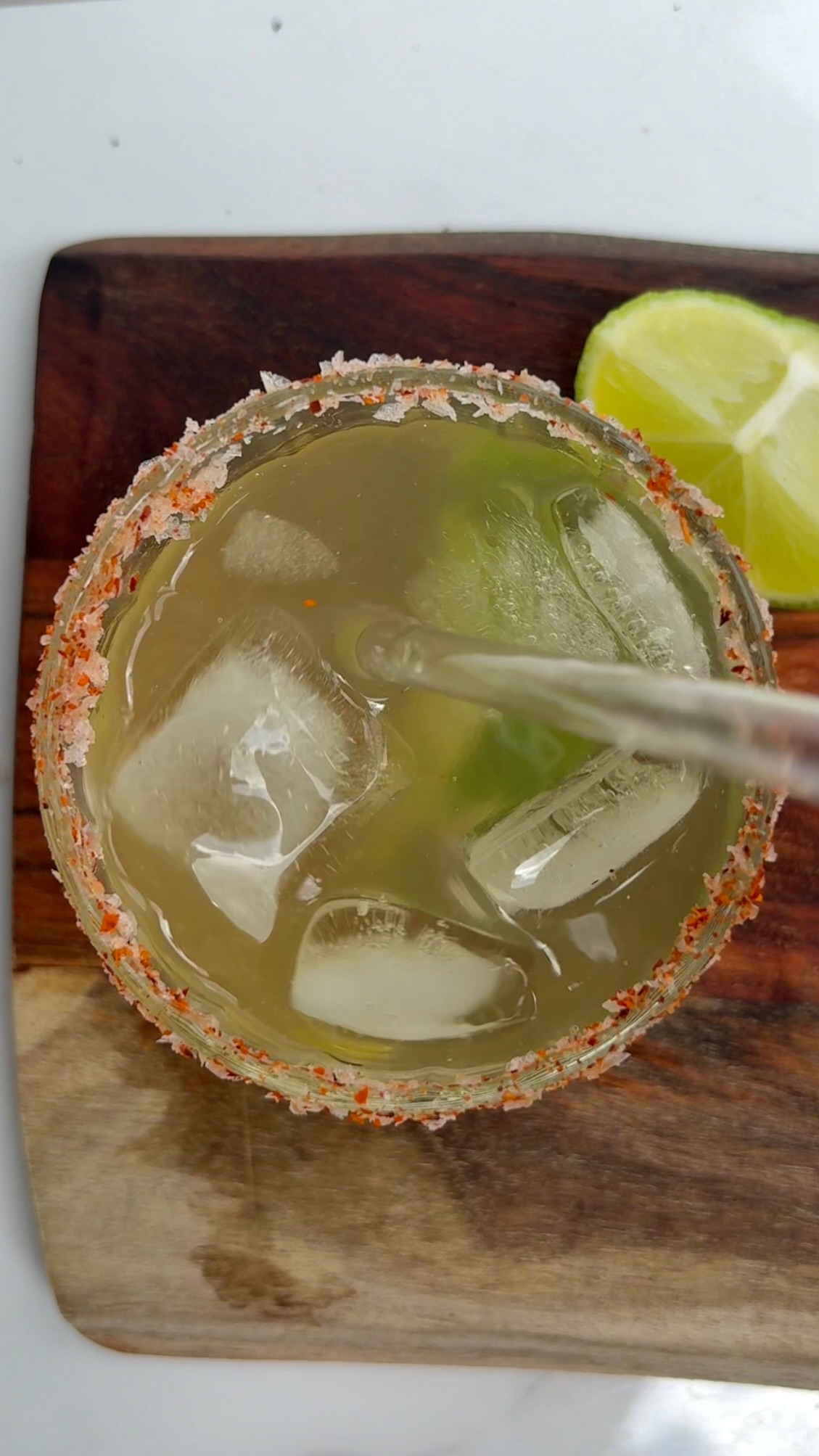 A close-up view of a cocktail in a glass with a salted rim, ice cubes, and a slice of lime on a wooden surface.