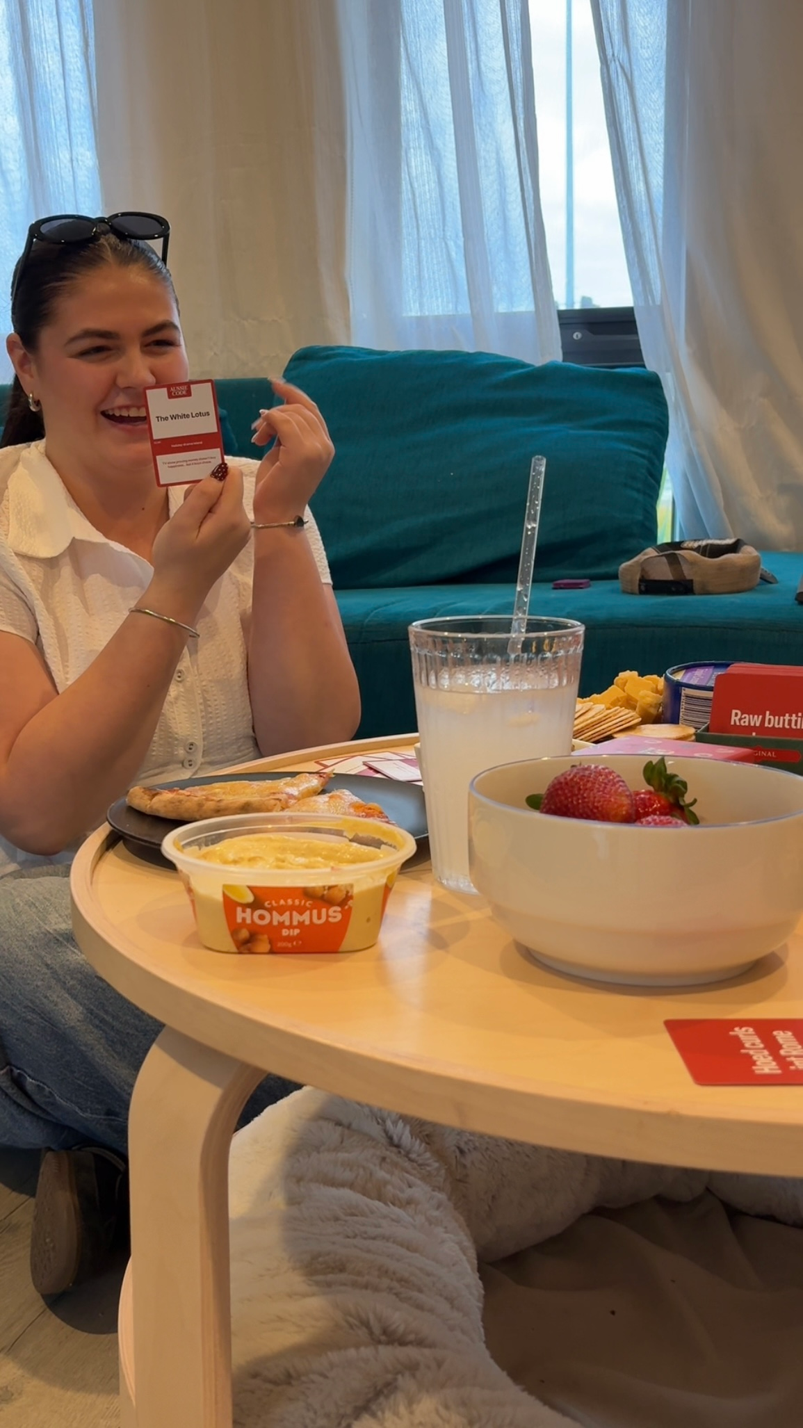 A young woman sitting at a table, smiling and holding a card while enjoying a meal that includes hummus, fruit, and snacks.