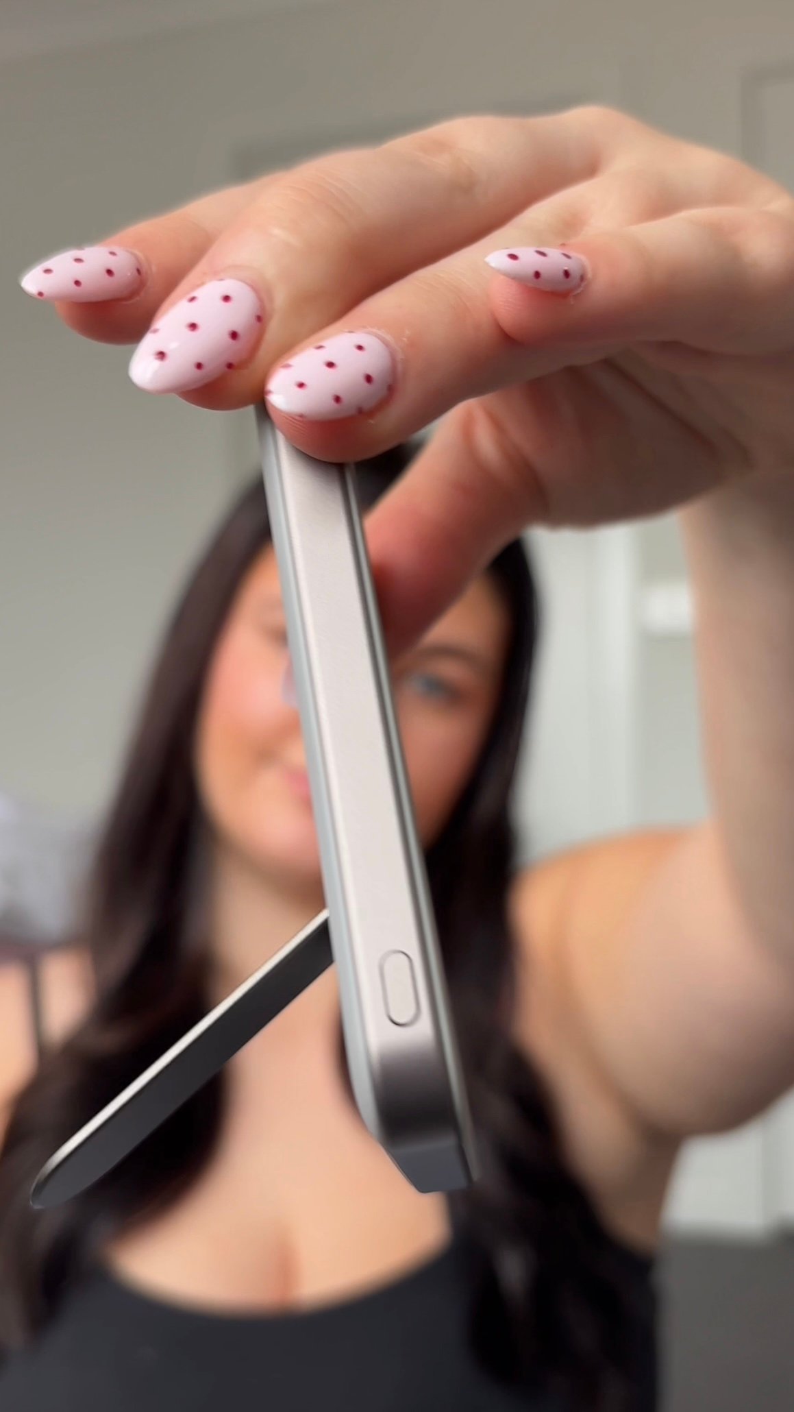 A close-up of a woman holding a smartphone with a sleek design, showcasing her manicured nails with pink polish and red dots.