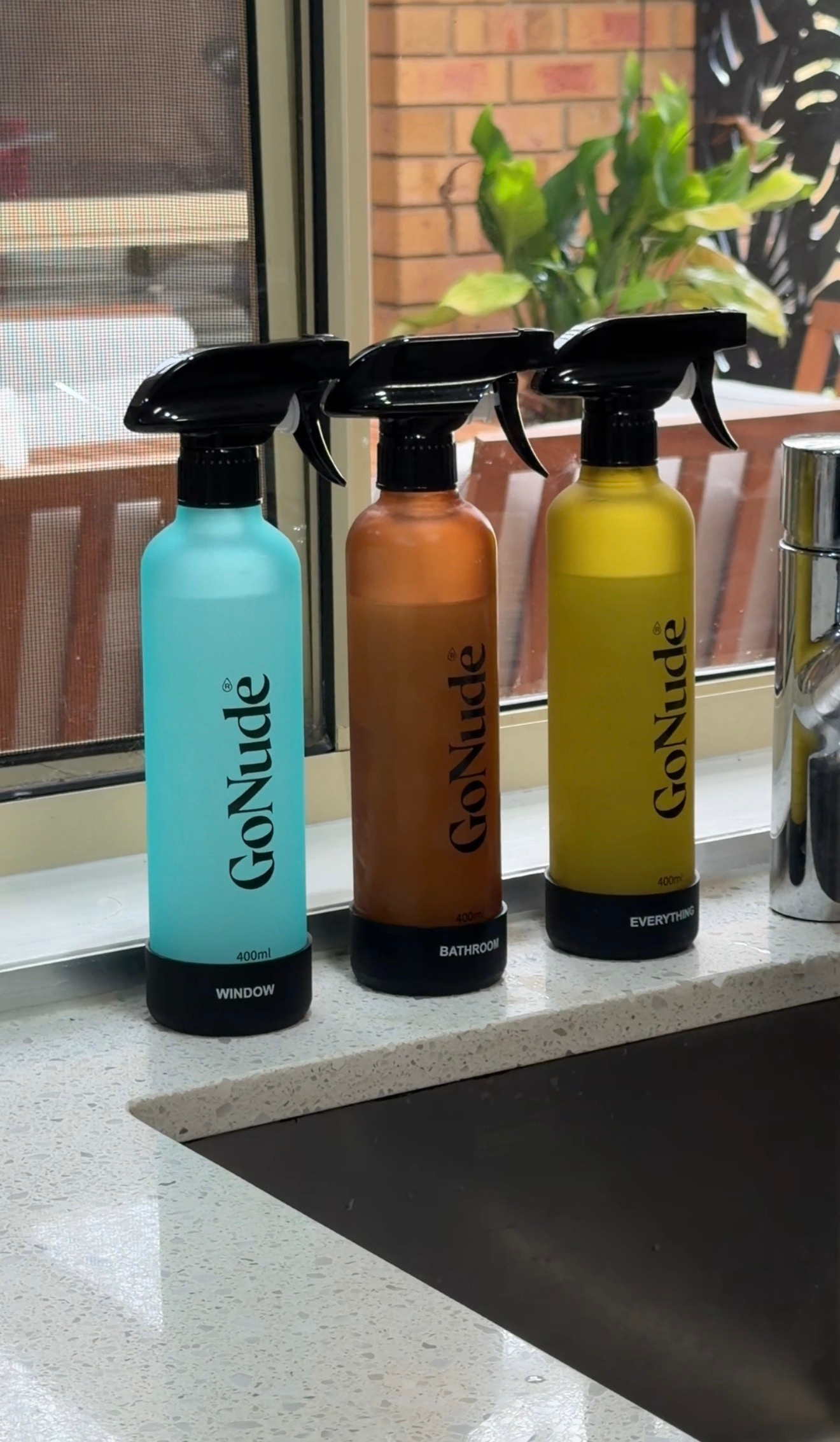 Three cleaning spray bottles in different colors arranged on a kitchen counter near a sink, labeled for window, bathroom, and general use.