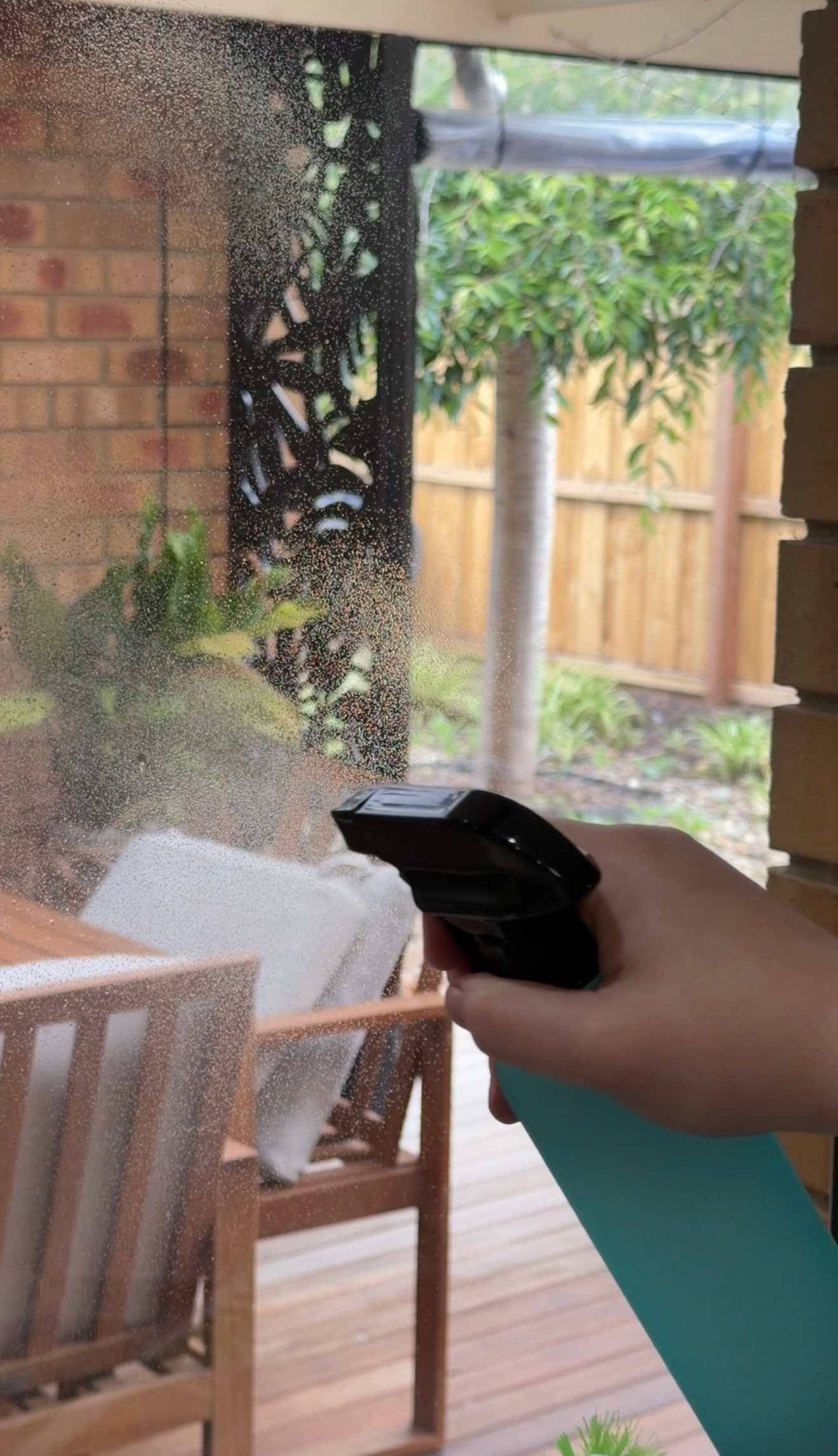 A hand holding a spray bottle, cleaning a glass window with a blurred outdoor setting in the background.