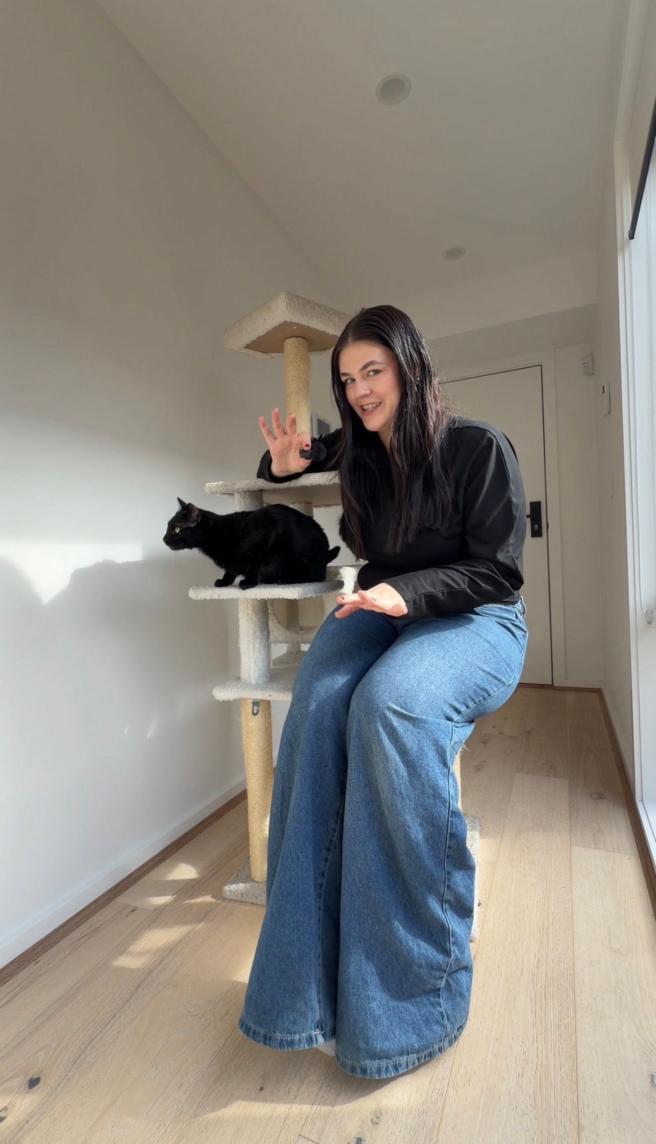 A person sitting on the floor next to a cat on a scratching post, gesturing playfully.