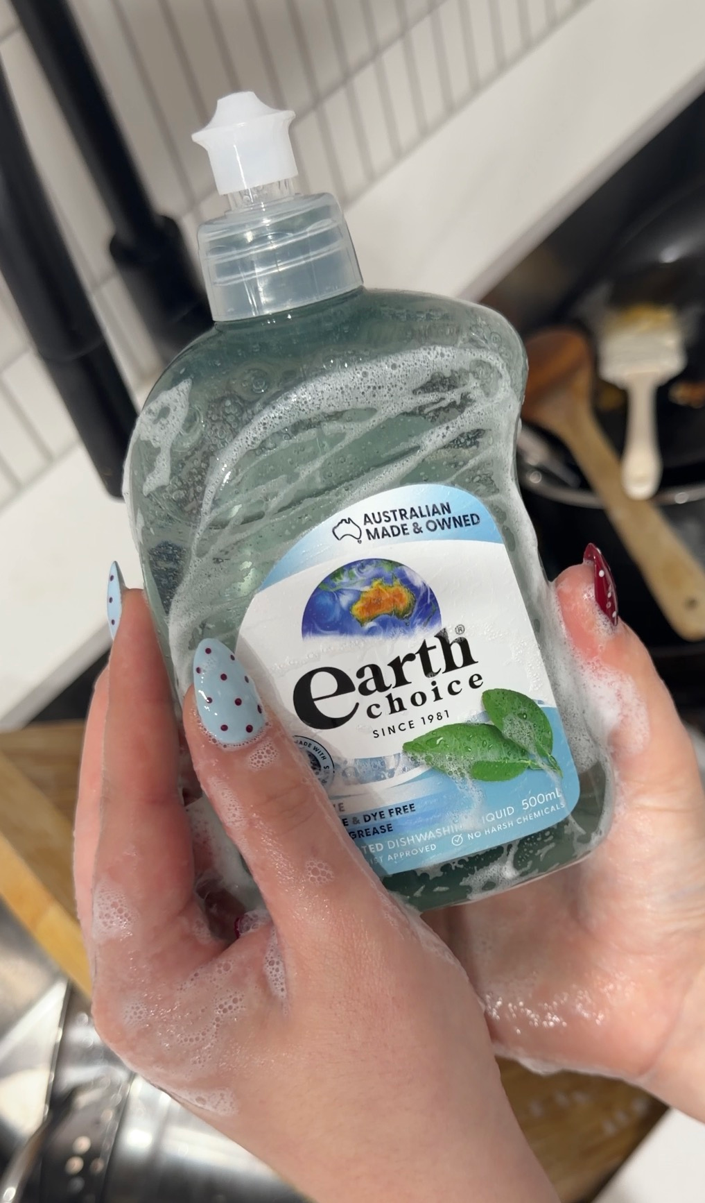 Hands holding a bottle of Earth Choice dishwashing liquid with bubbles, set against a kitchen background.