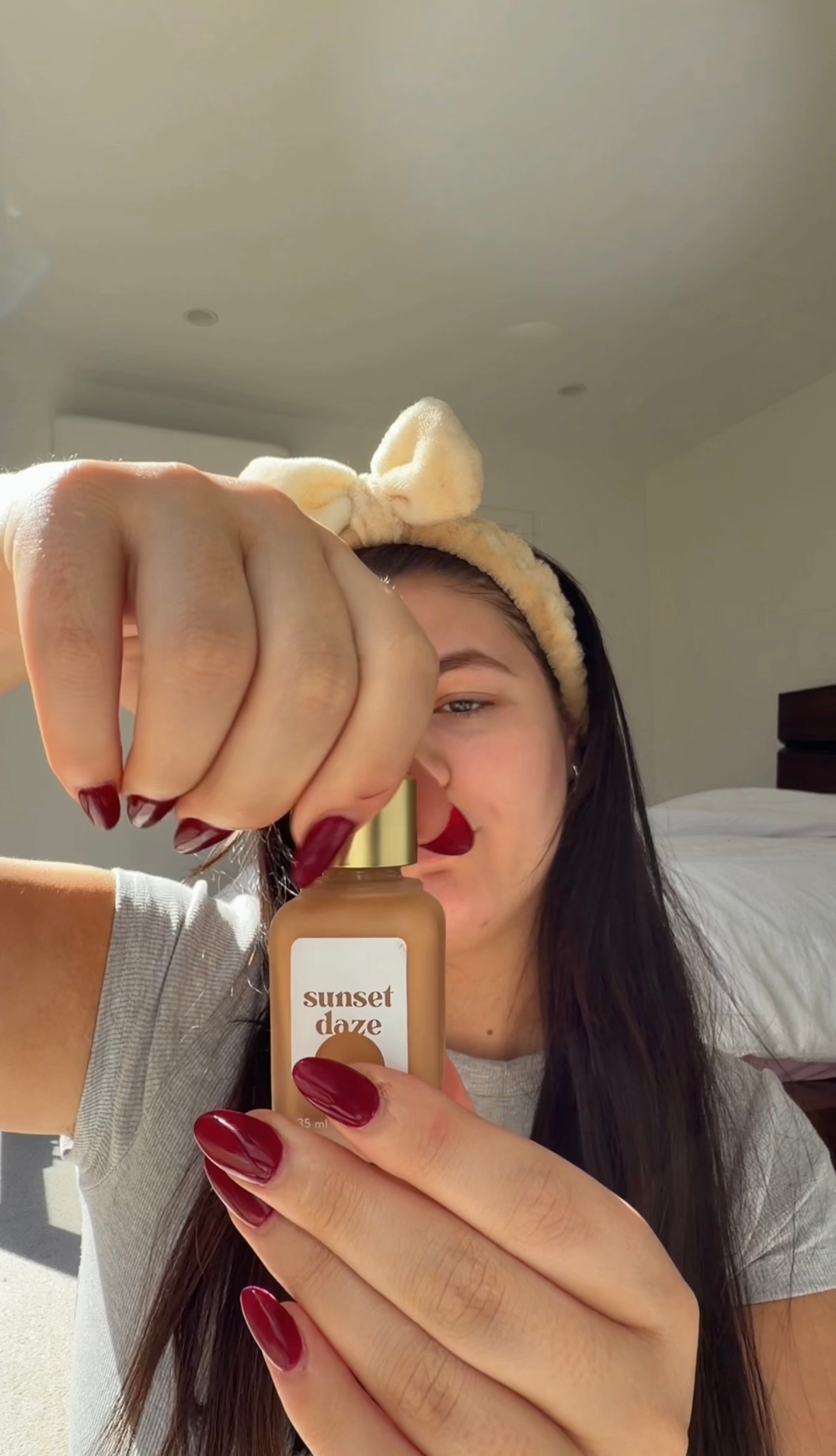 A person with long dark hair and a headband is holding a bottle of 'sunset dive' product, showcasing it with manicured red nails.