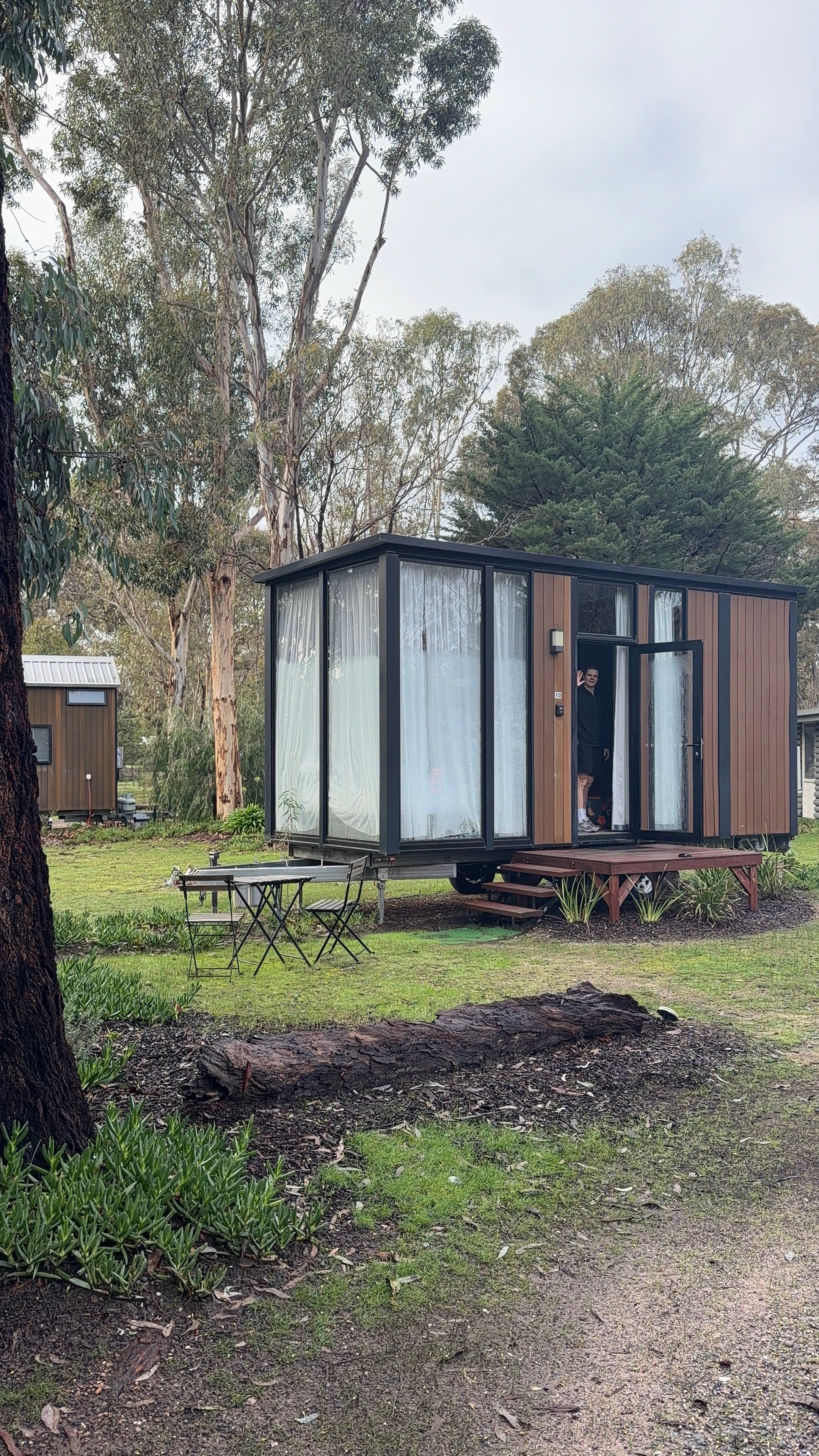 A modern tiny house with large windows, set in a natural landscape surrounded by trees. A person is visible inside, looking out.