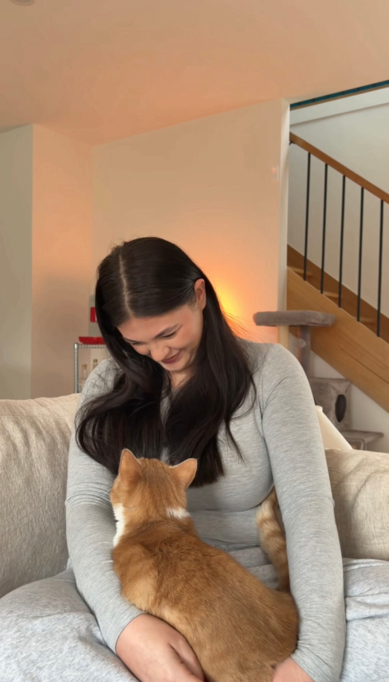 A woman with long dark hair smiles while sitting on a couch, gently interacting with a ginger cat in her lap.
