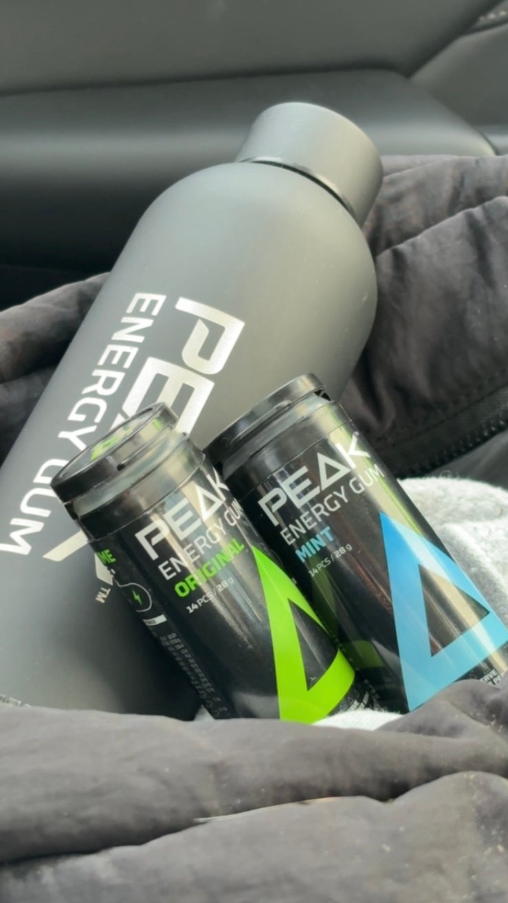 A black water bottle and two containers of Peak Energy Gum, one labeled orange and the other labeled mint, resting on a soft surface.