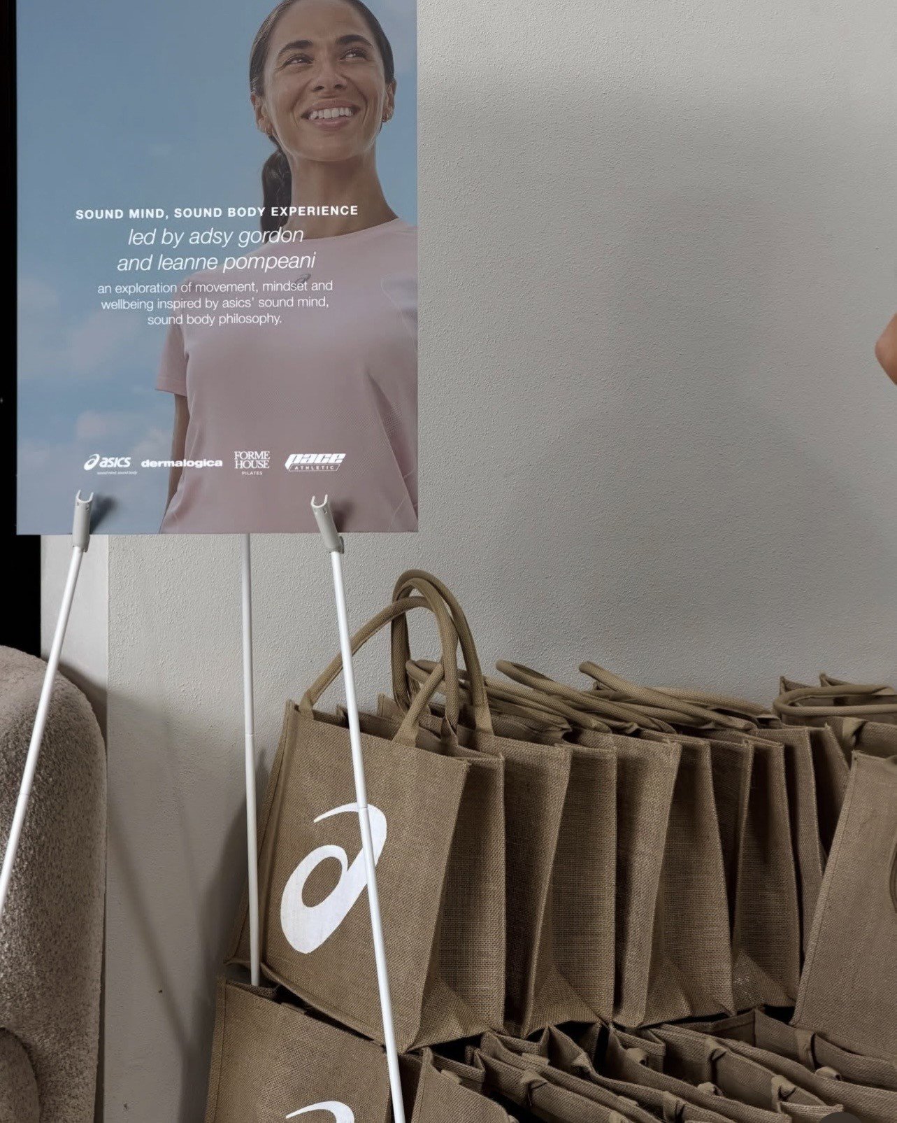A woman smiling in front of a promotional poster for a wellness experience, with a stack of burlap bags in the foreground.