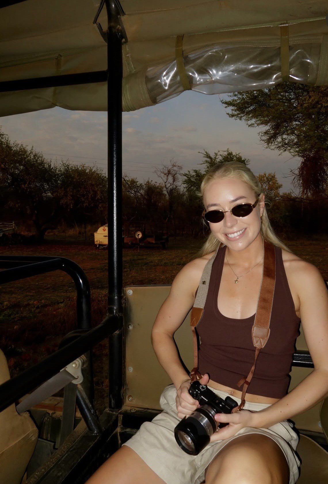 A young woman sitting in a safari vehicle, wearing sunglasses and holding a camera, with a natural landscape in the background.