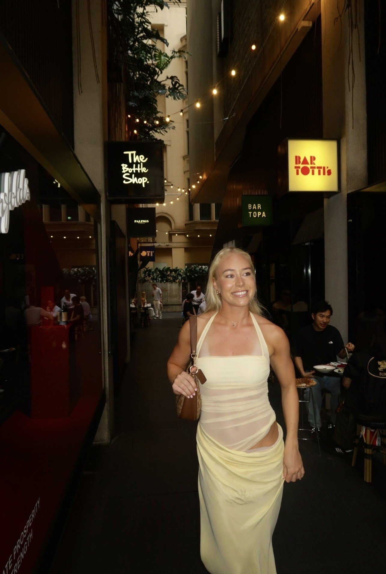 A woman in a sheer yellow dress walks confidently through a lively urban alleyway filled with shops and diners.