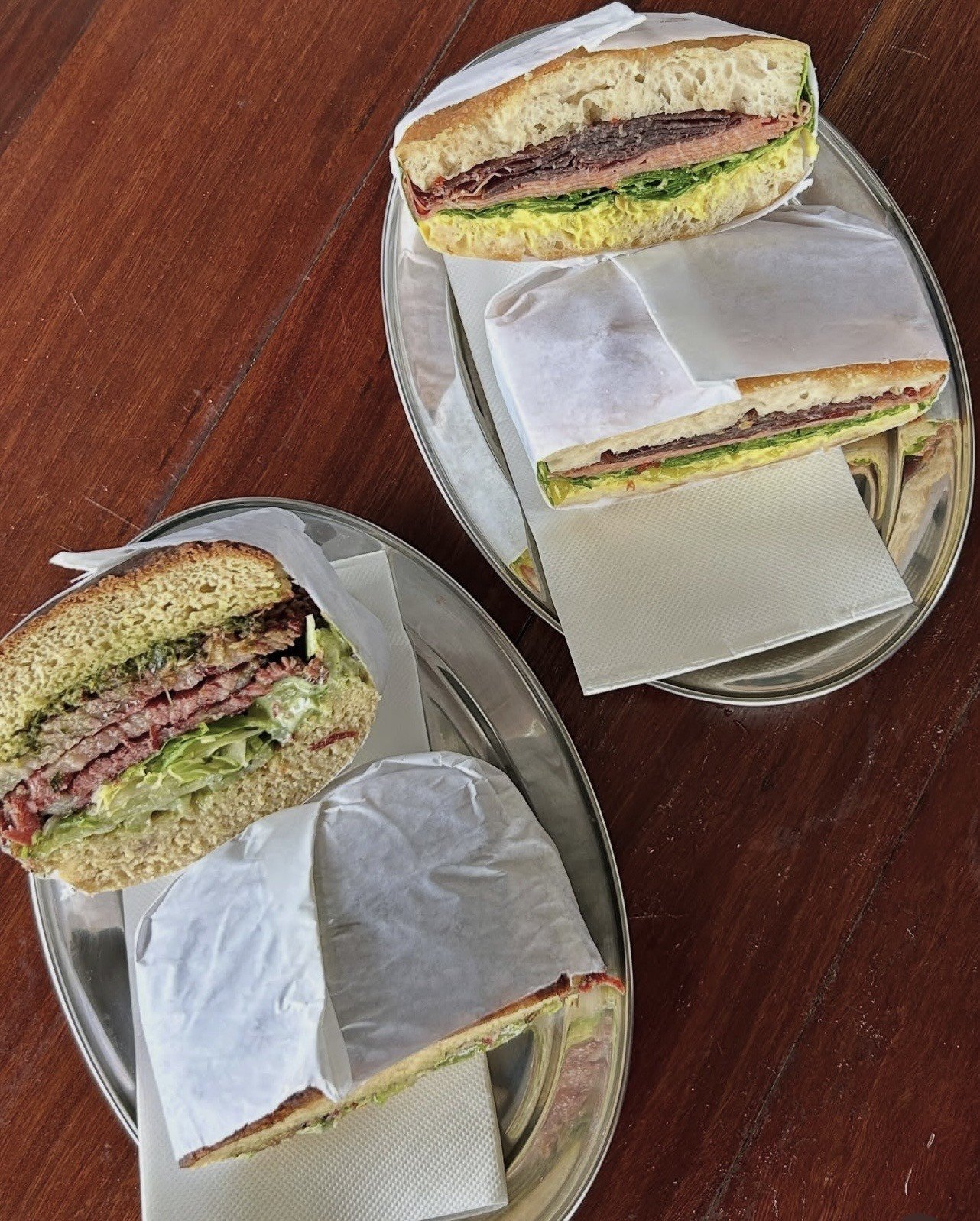 Two sandwiches on silver trays, wrapped in paper, showcasing layers of meat, lettuce, and other fillings.