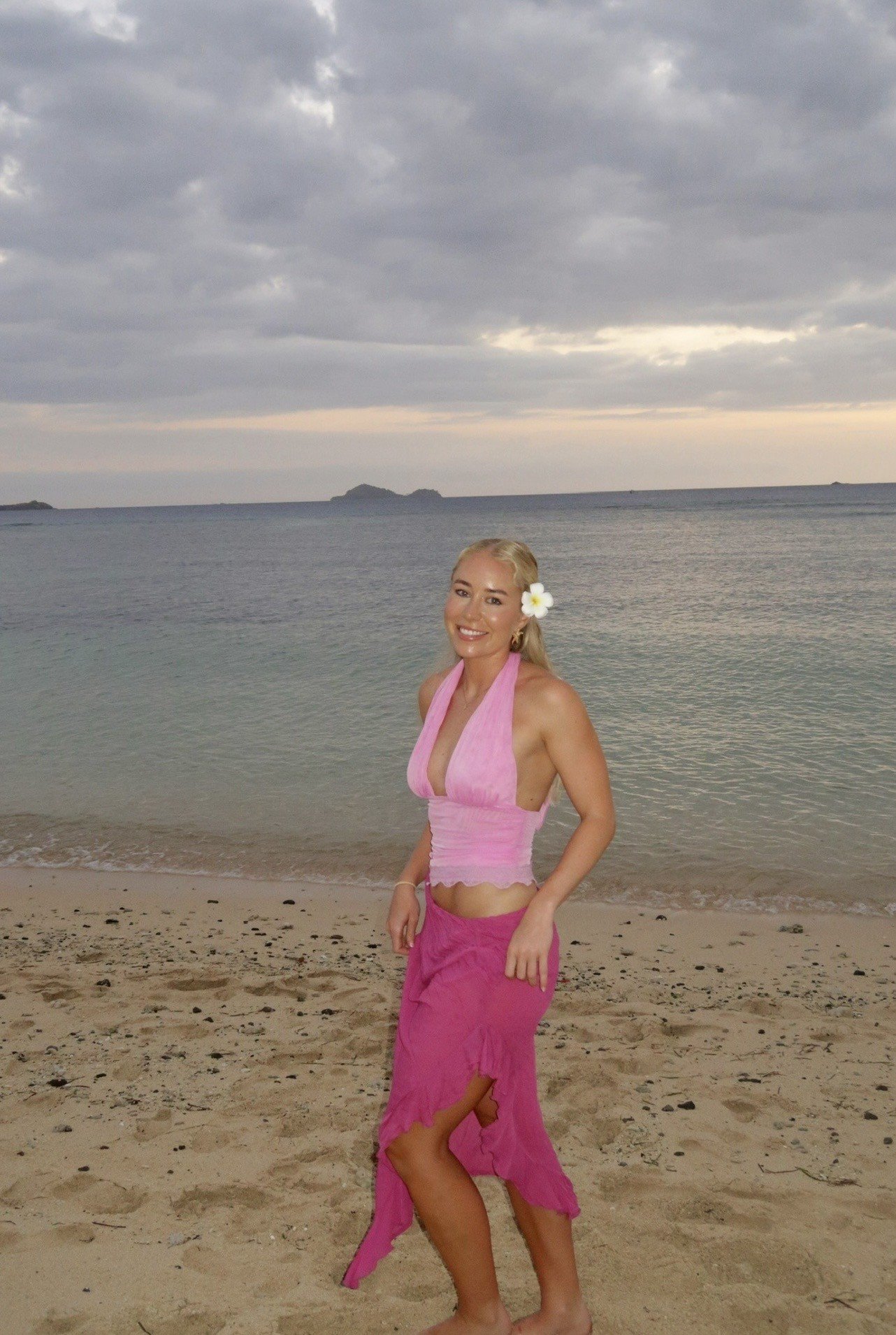 A woman in a pink outfit stands on a beach with a serene ocean backdrop and cloudy sky.