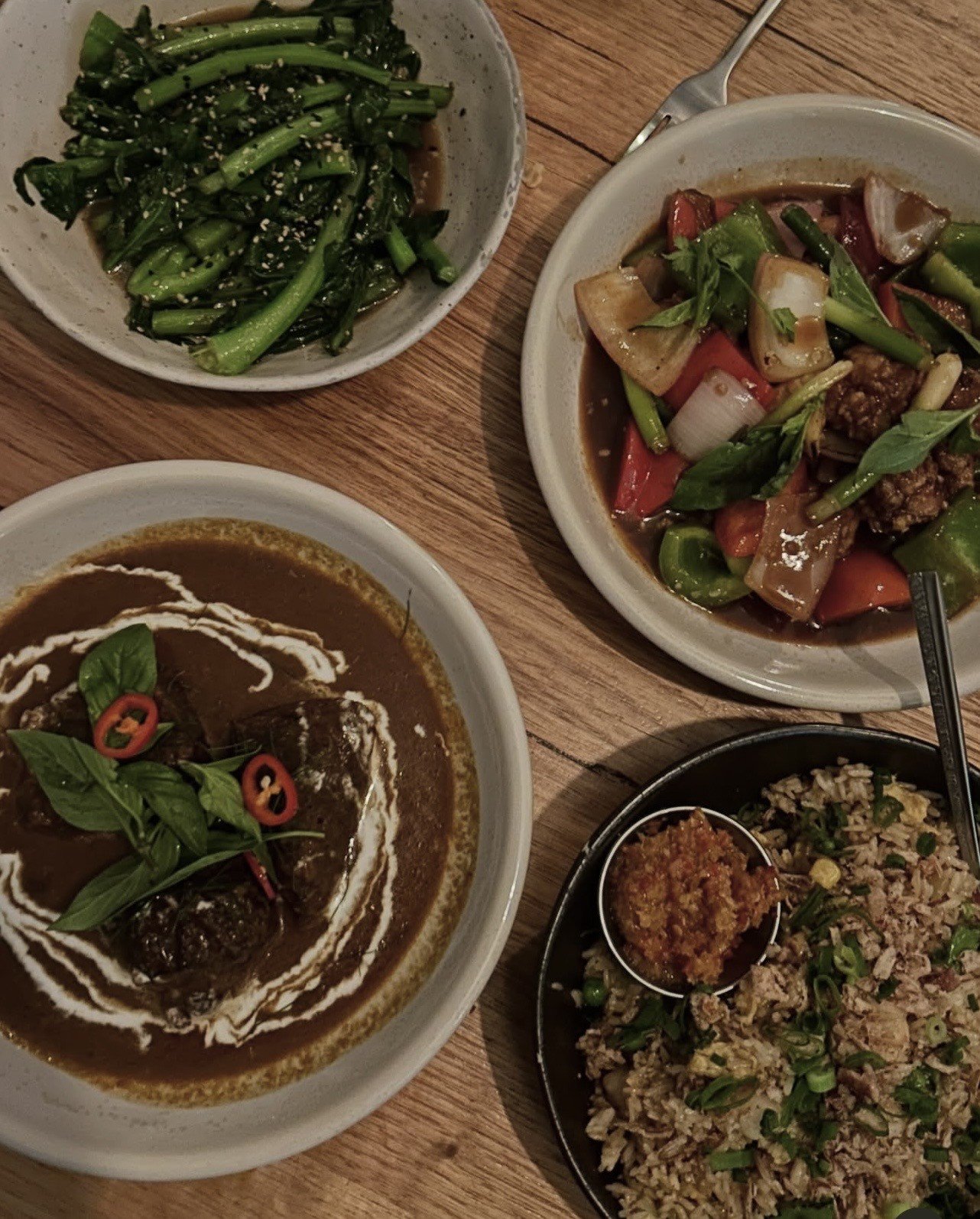 A variety of Asian dishes arranged on a wooden table, including a curry with herbs, stir-fried greens, a sweet and sour dish, and fried rice.