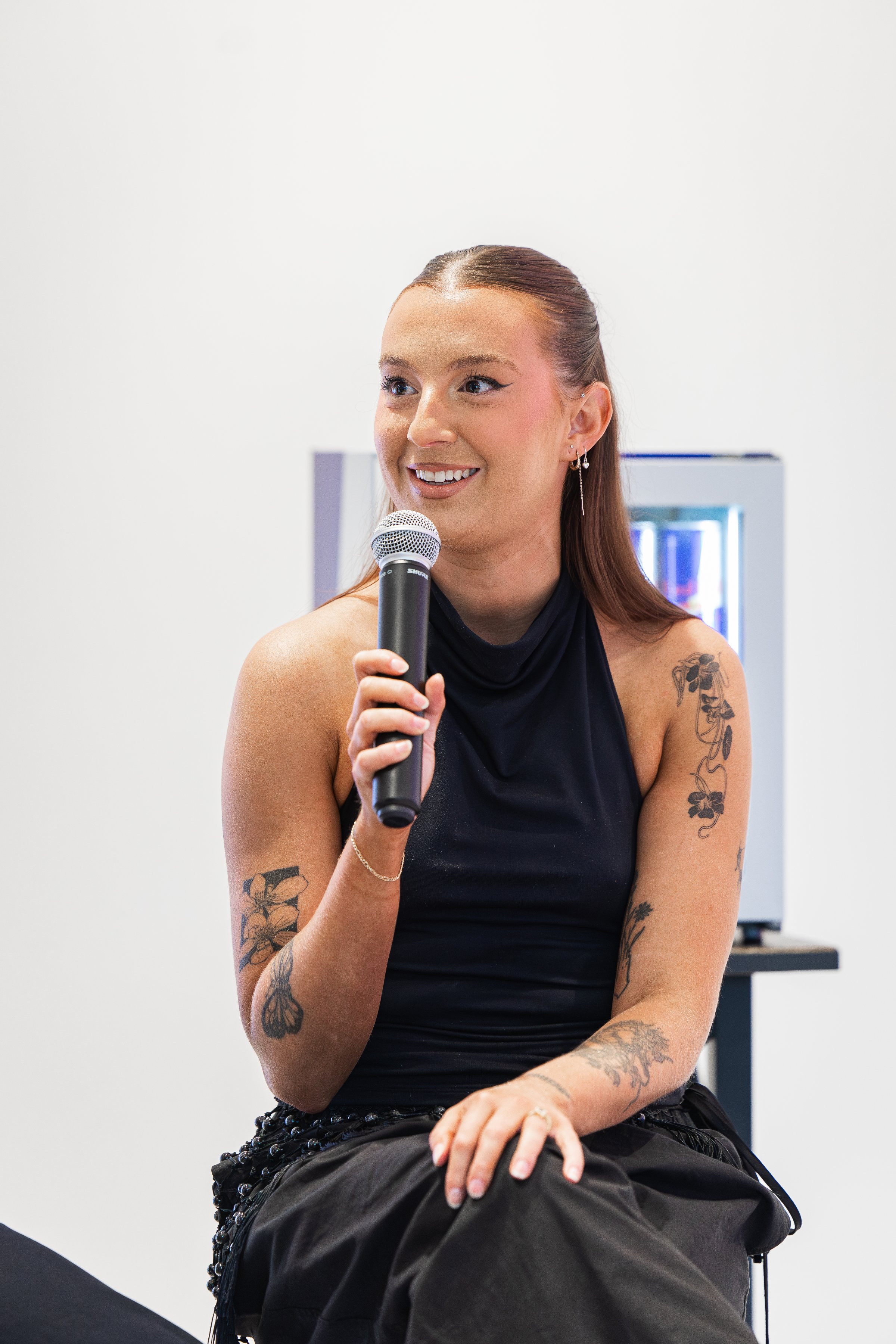 A woman with tattoos smiles while holding a microphone, seated in front of a minimalist background.