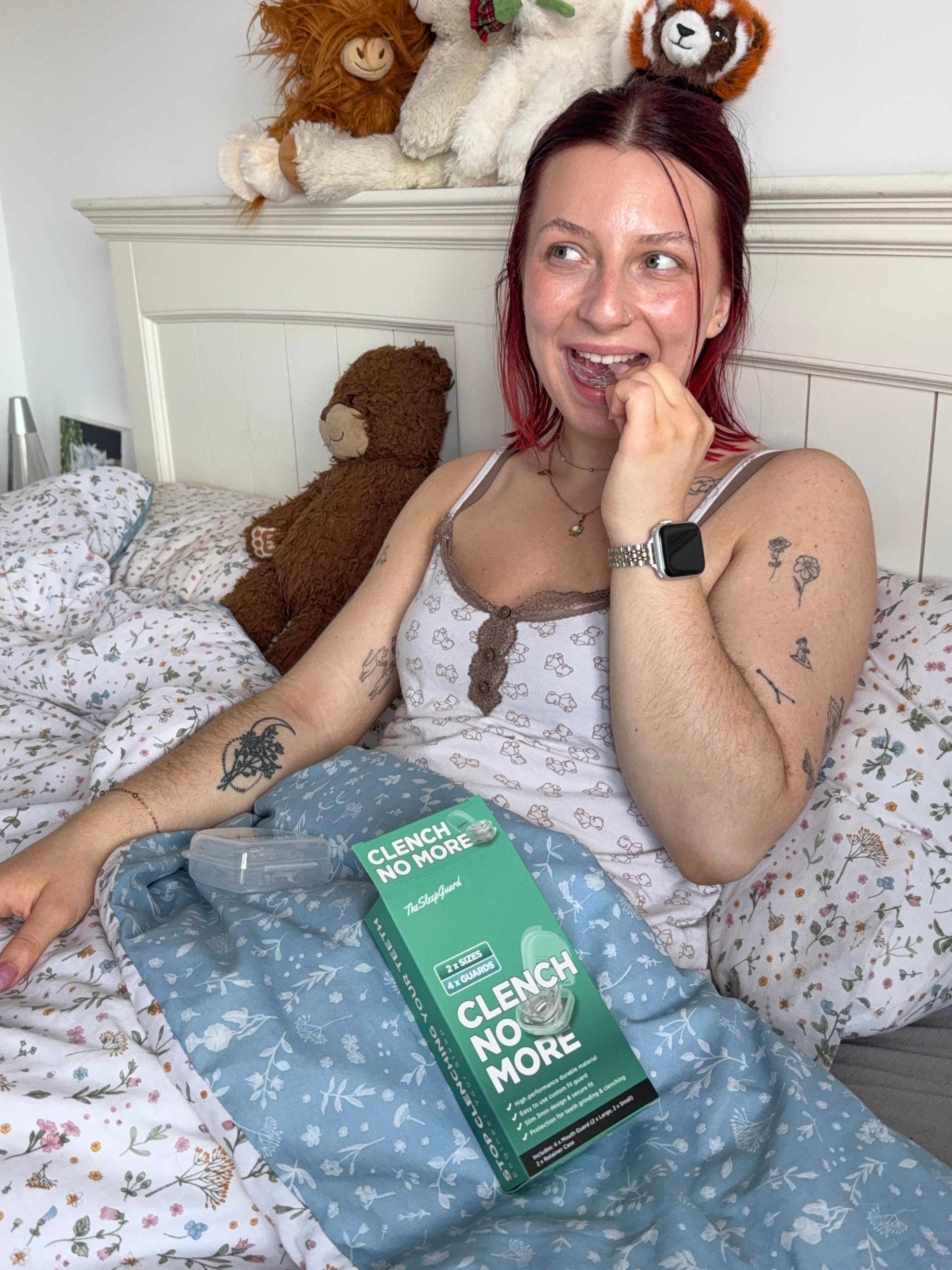 A young woman sitting on a bed with a playful expression, holding a product box labeled 'CLENCH NO MORE'. She has tattoos and is surrounded by stuffed animals.