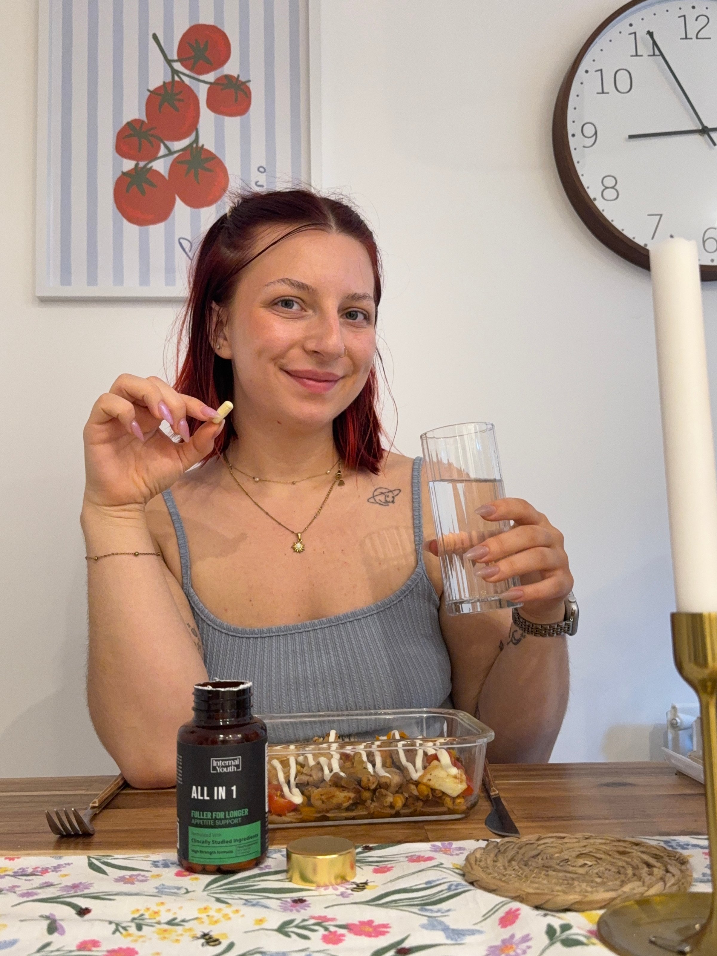 A woman sitting at a table, smiling while holding a glass of water and a supplement capsule, with a meal in front of her and a decorative clock on the wall.