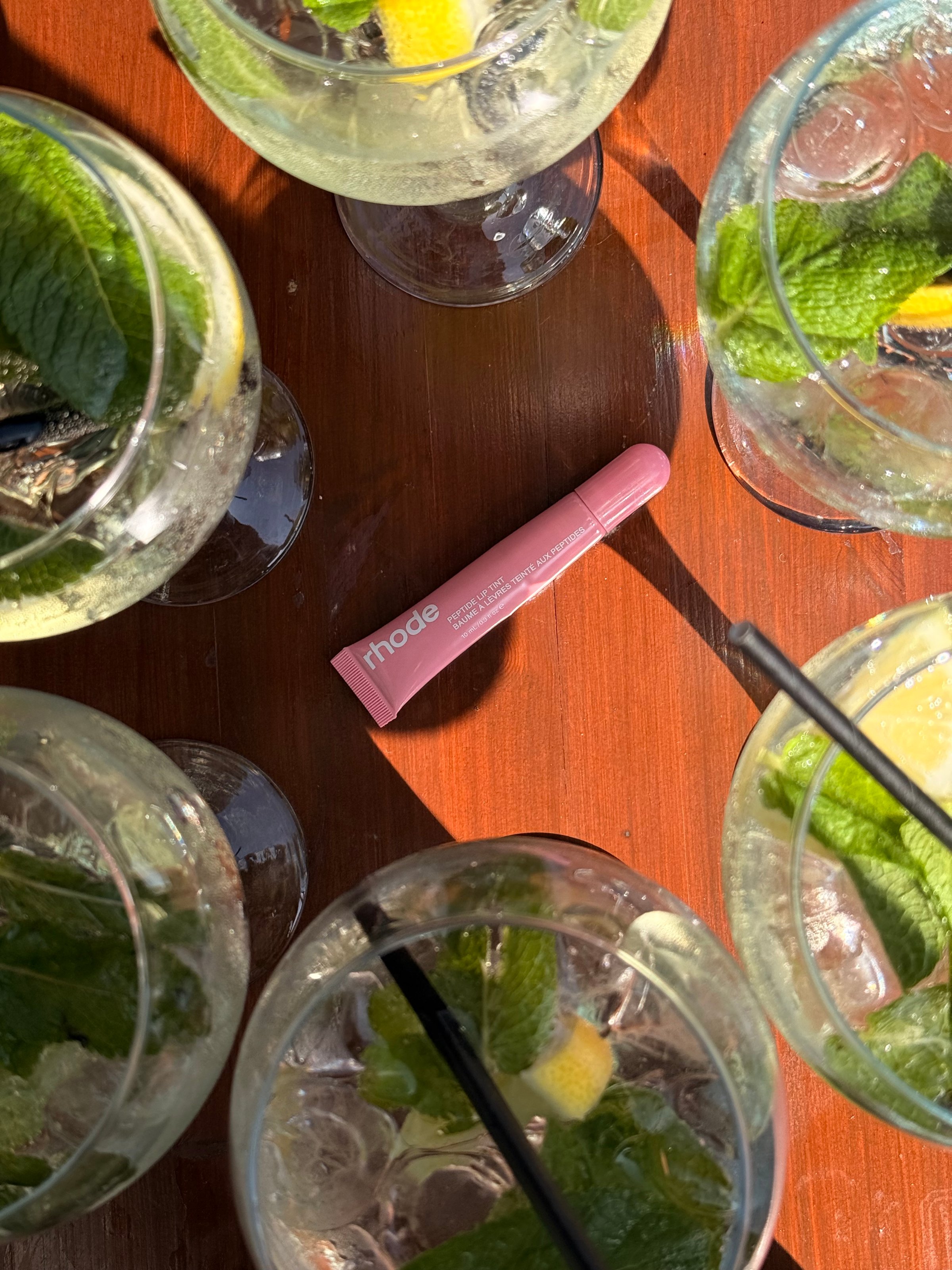 A top-down view of several cocktails with mint and lemon, surrounding a pink lip balm tube on a wooden surface.